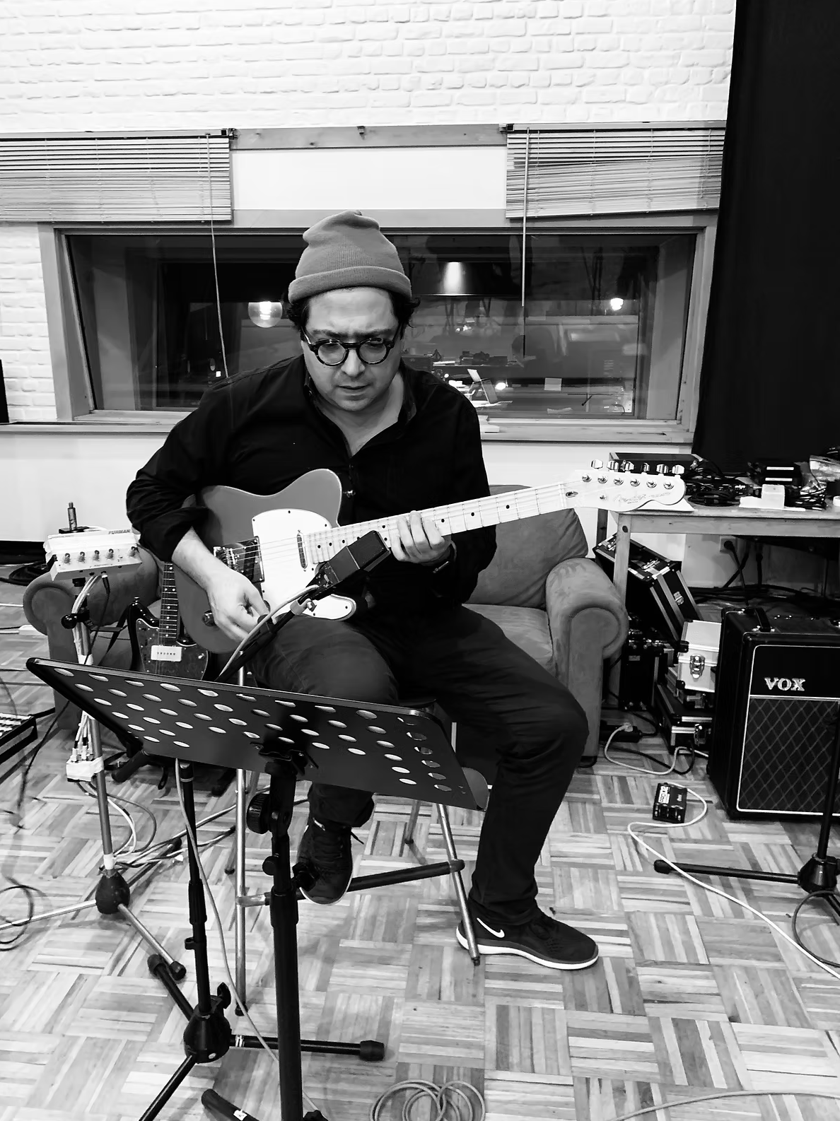 A man with glasses and a beanie playing an electric guitar in a recording studio.