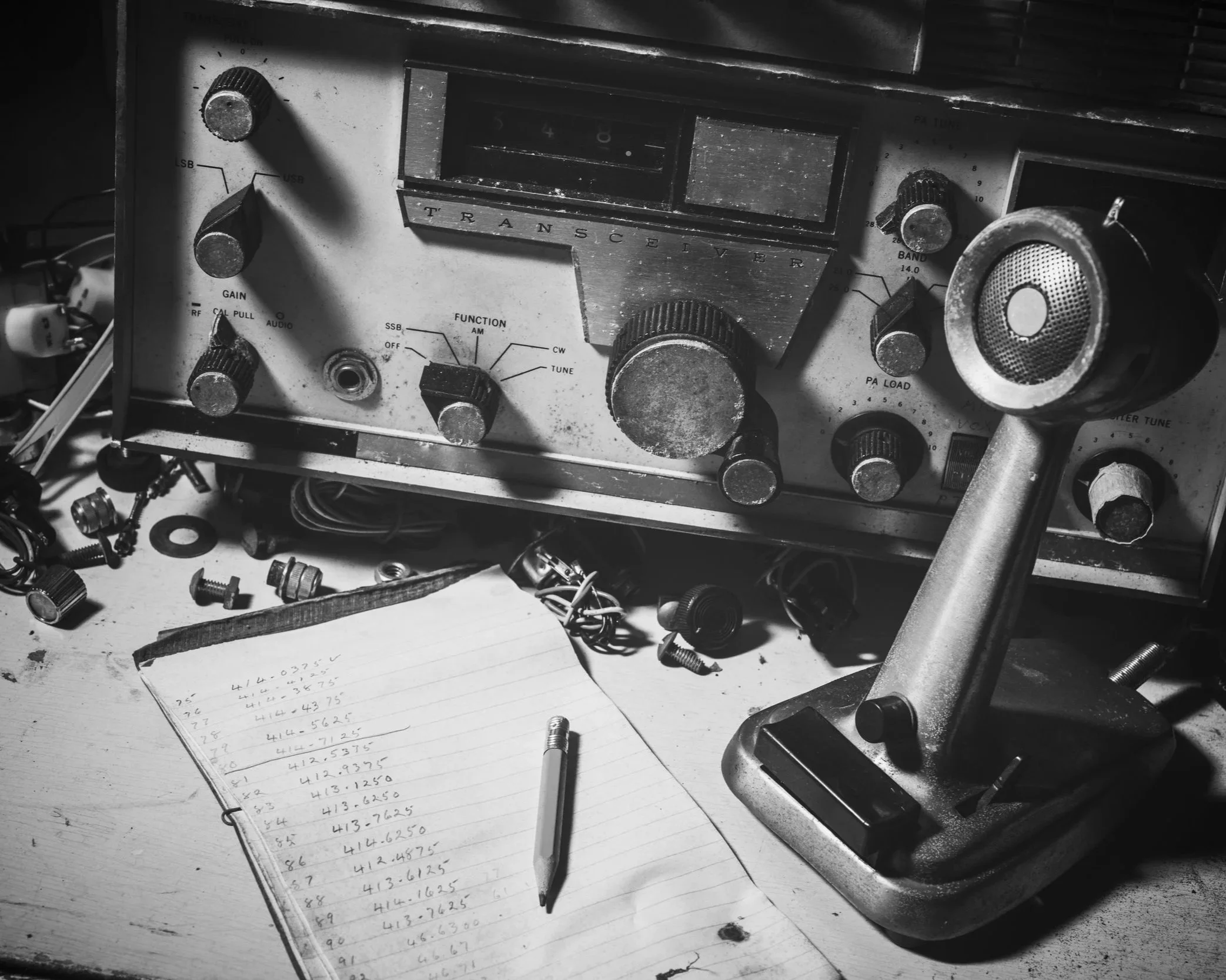 Black and white photo of vintage transceiver radio, a notebook with handwritten numbers, and scattered electronic components on a desk.