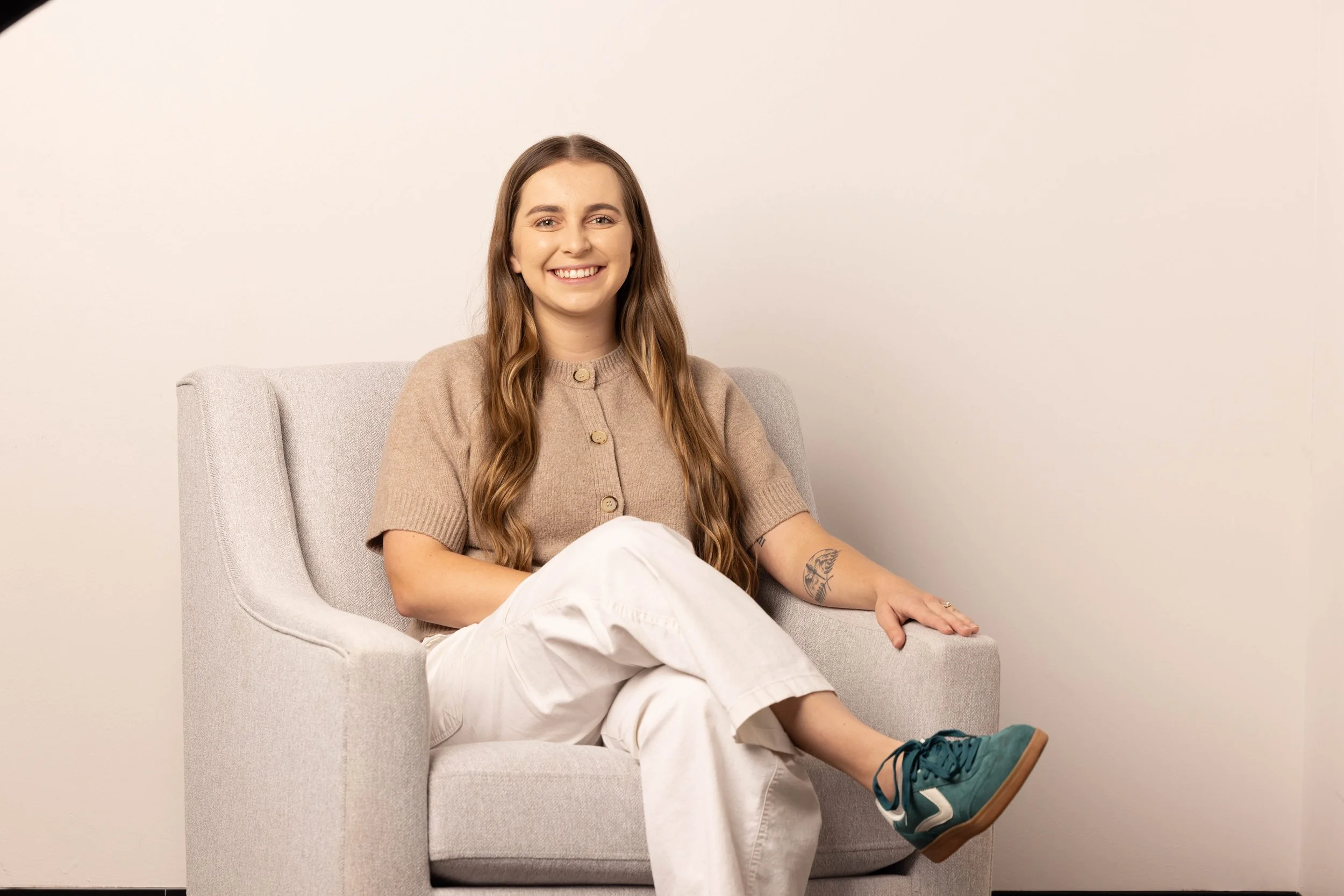A young woman with long wavy brown hair, smiling, sitting on a light gray armchair in front of a plain beige wall. She is wearing a beige cardigan, white pants, and teal sneakers, with a tattoo of a feather on her left forearm.