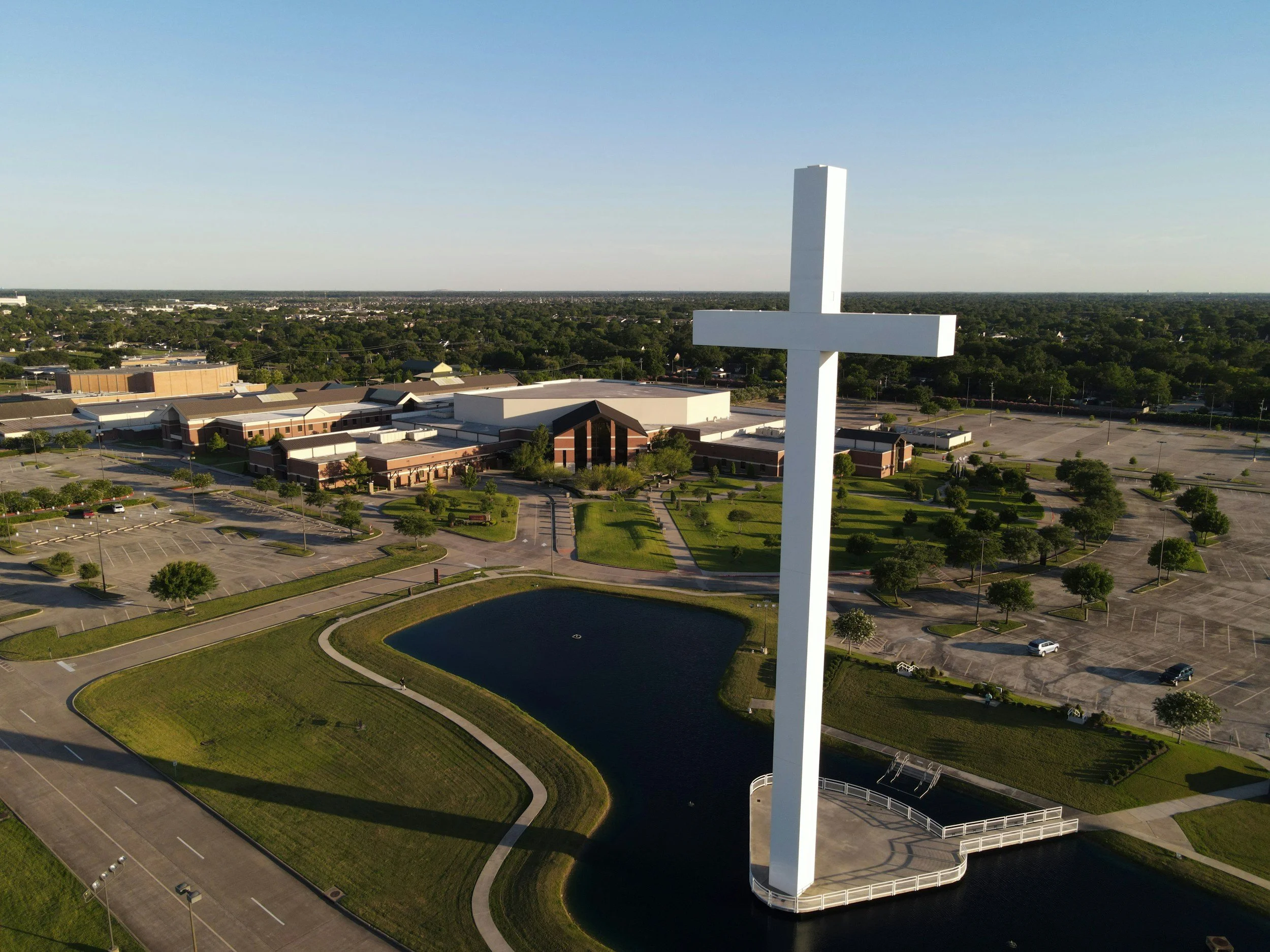 A large white cross sculpture standing on a platform over a small pond, surrounded by a parking lot, trees, and a church building in the background under a clear blue sky.