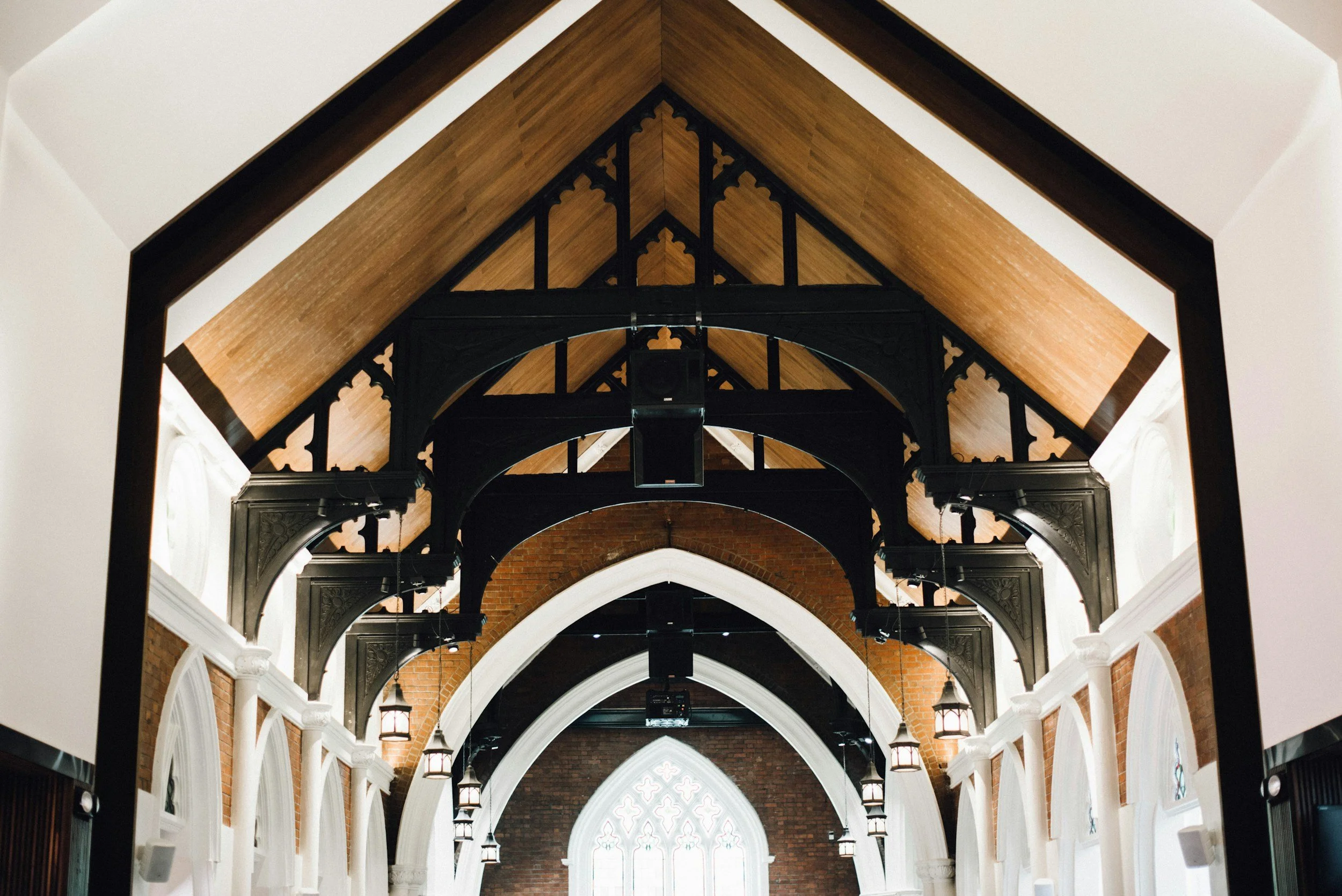 Interior of a building with gothic-style arches, wooden ceiling, brick walls, and hanging lantern lights.