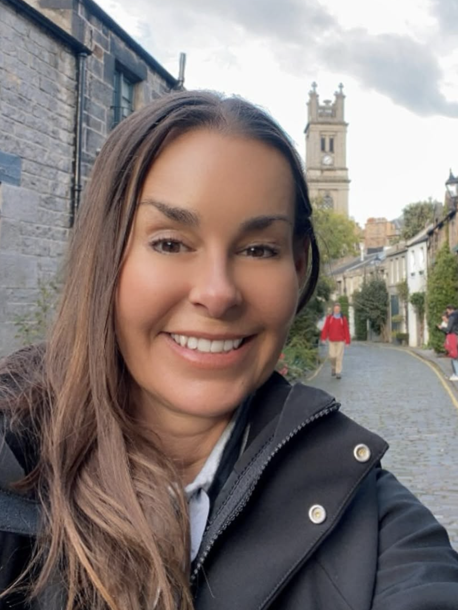 A smiling woman taking a selfie outdoors on a cobblestone street with a church tower in the background.