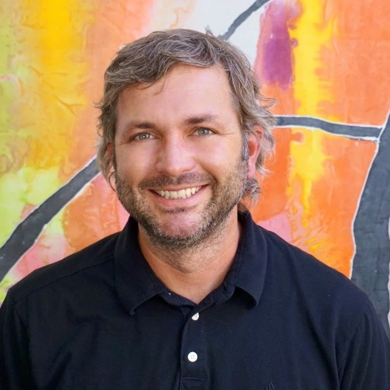 A smiling man with gray hair and a beard, wearing a black shirt, standing in front of a colorful abstract background.