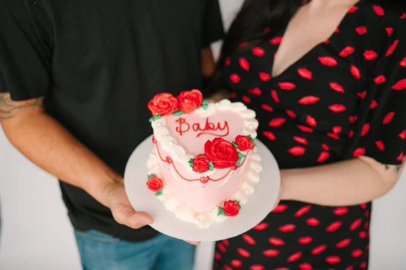 Close-up of a person's foot wearing a black strappy high-heeled sandal, dangling over a plate of sliced cake, with a backdrop of red roses.
