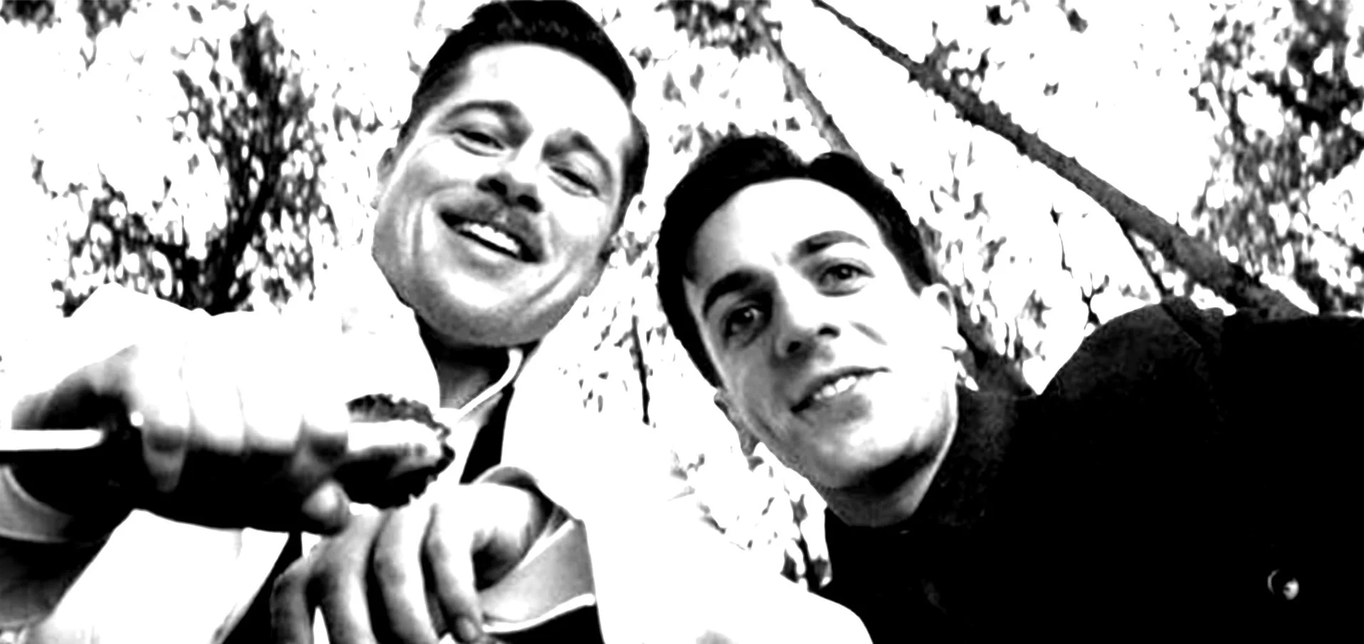 Black and white photograph of two young men smiling for a selfie outdoors, with trees and sky visible in the background.