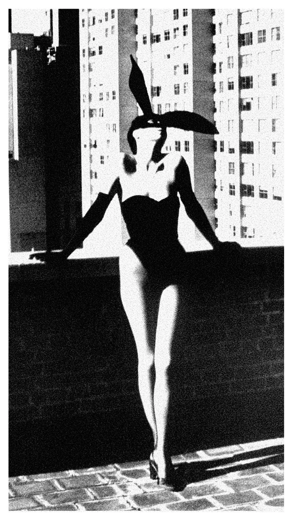 A black-and-white photo of a woman in a one-piece swimsuit and bunny ears leaning on a ledge with an urban building and windows in the background.