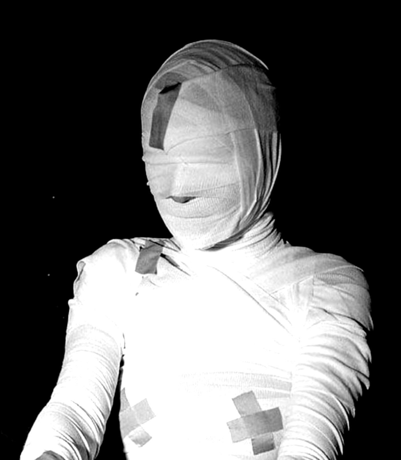 Black and white photo of a person wrapped entirely in bandages, with their face covered, standing against a dark background.