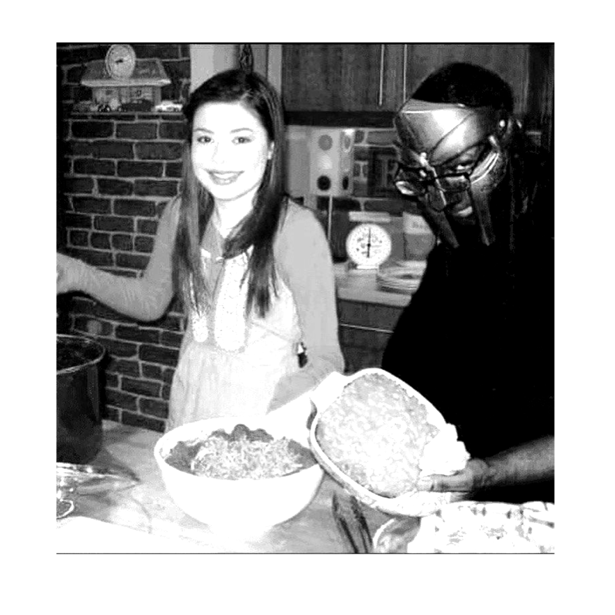 Two women in a kitchen, one smiling at the camera and the other wearing protective glasses, holding a large bowl of food. The kitchen has wooden cabinets, a brick wall, and various items on the countertop.