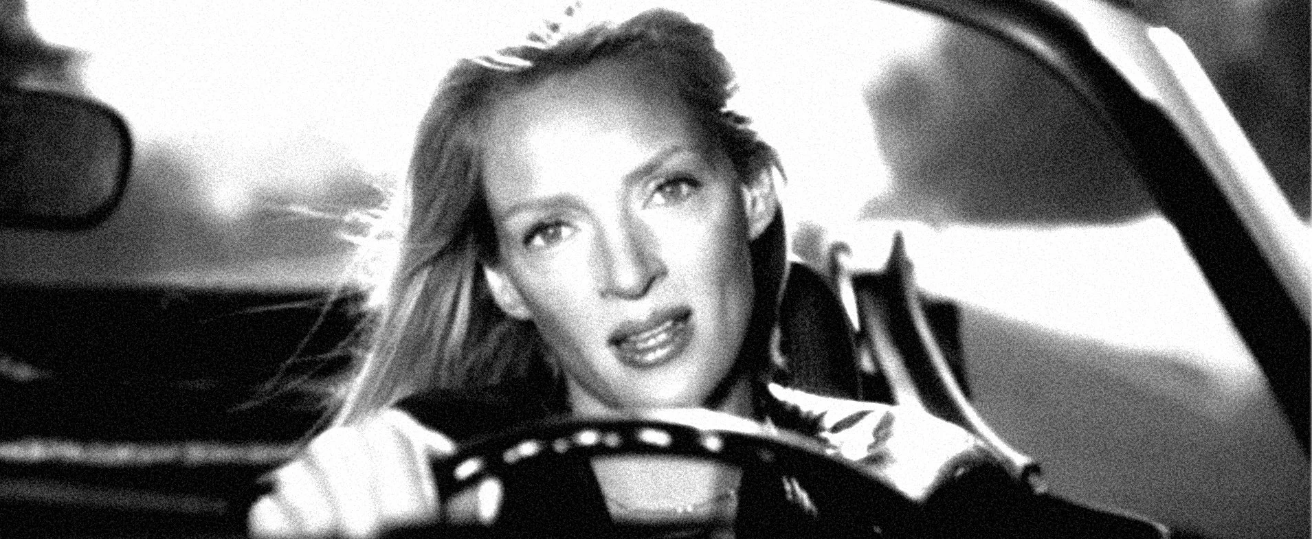 Black and white photo of a woman with long hair, sitting in a car, looking forward with an intense expression.