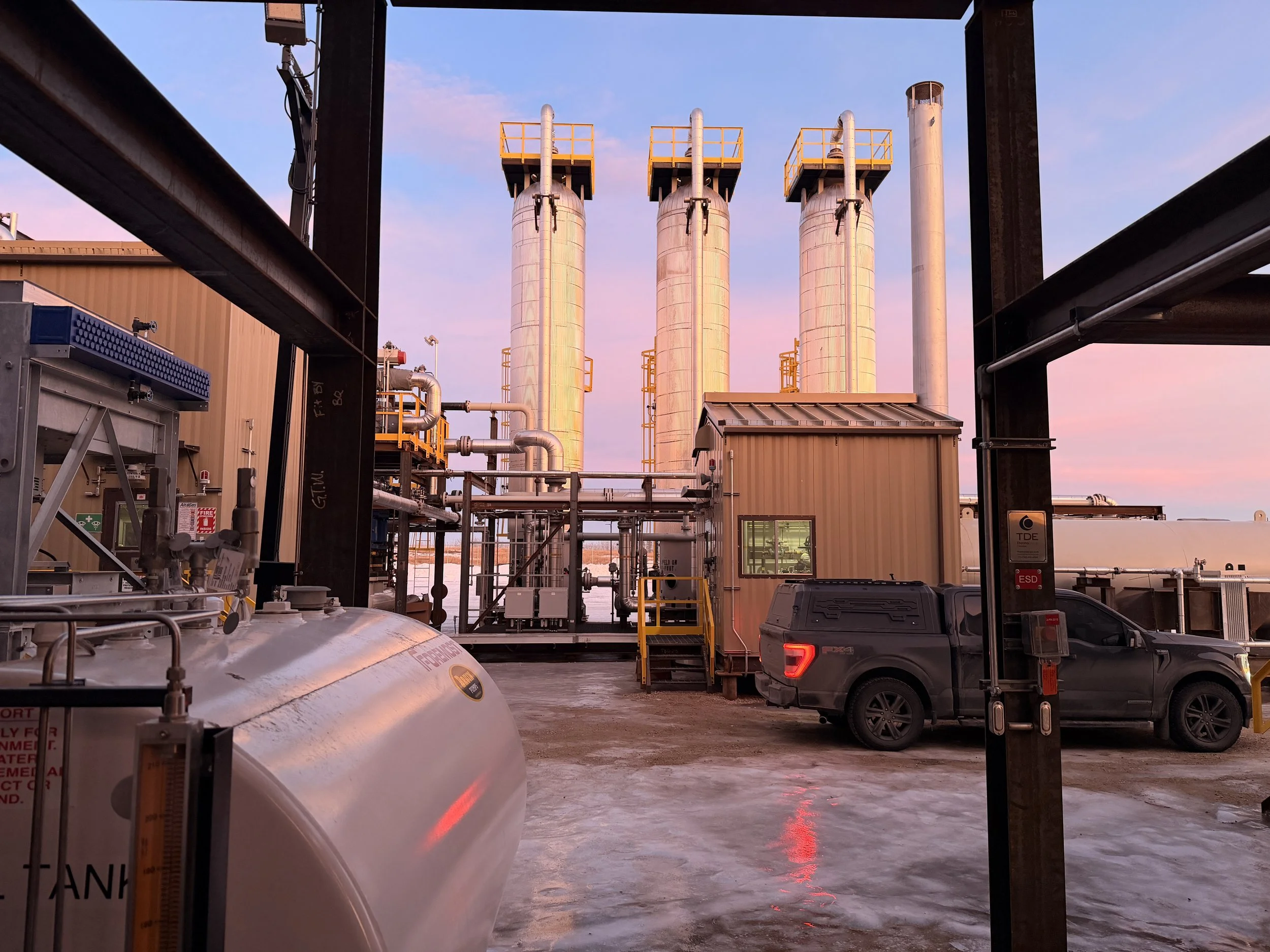 Industrial facility with large vertical tanks and pipes, a yellow and black vehicle, and a beige building under a pink and blue sky at sunset.