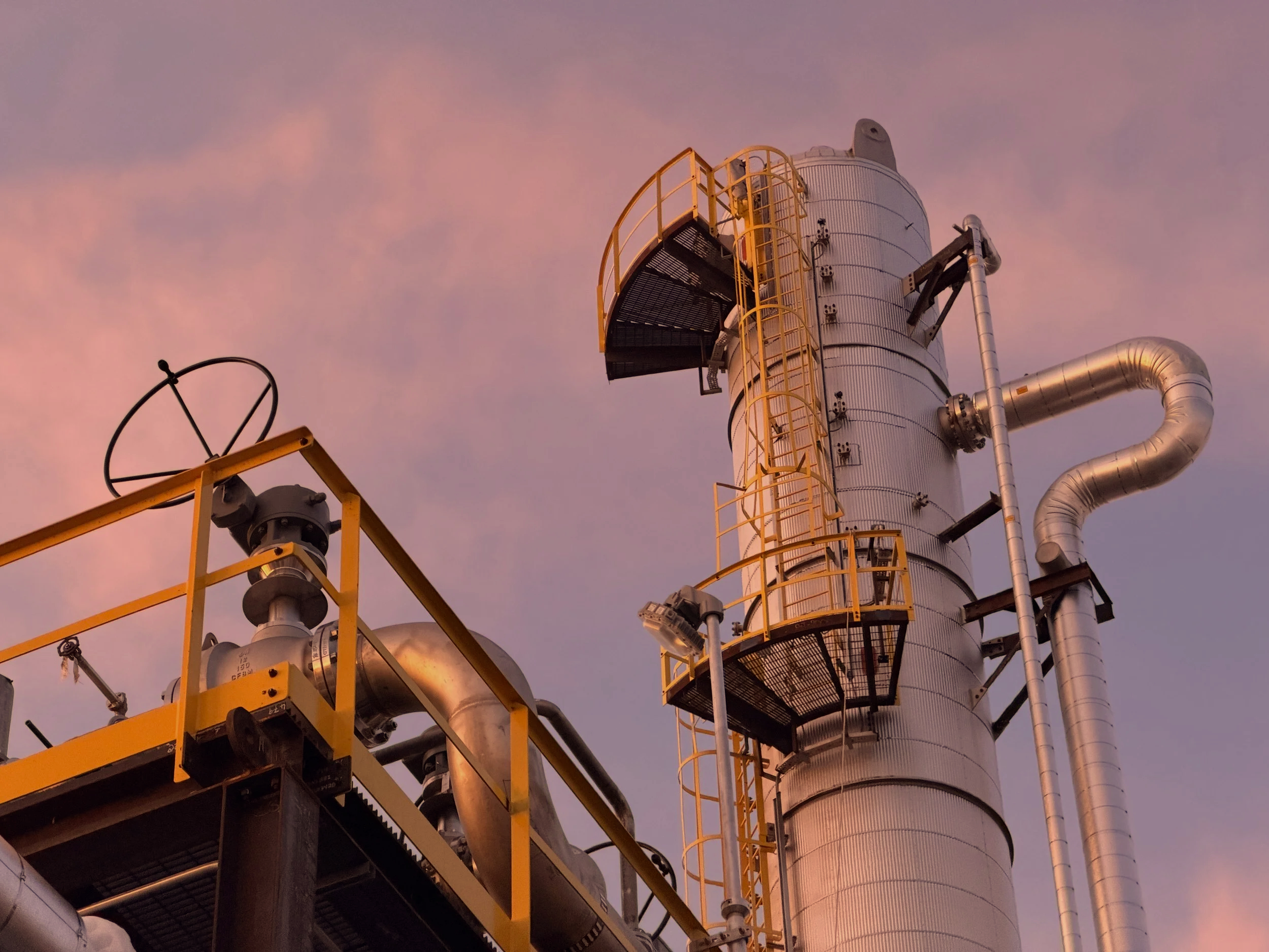 Industrial refinery with large metal pipes and a tall cylindrical tower against a pink and purple sky.