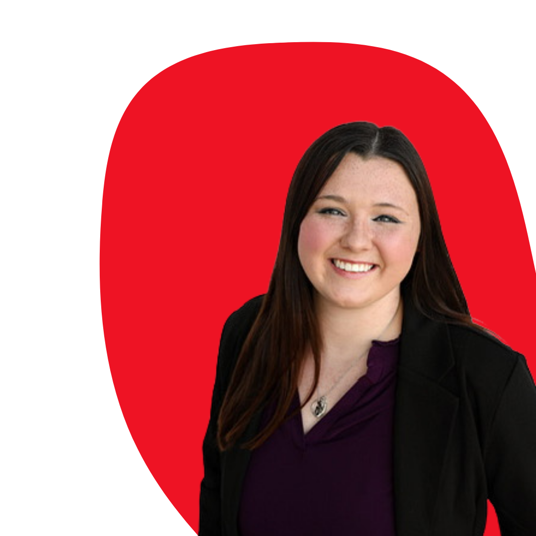 A woman with long brown hair smiling, wearing a black blazer and purple top, standing in front of a red background.