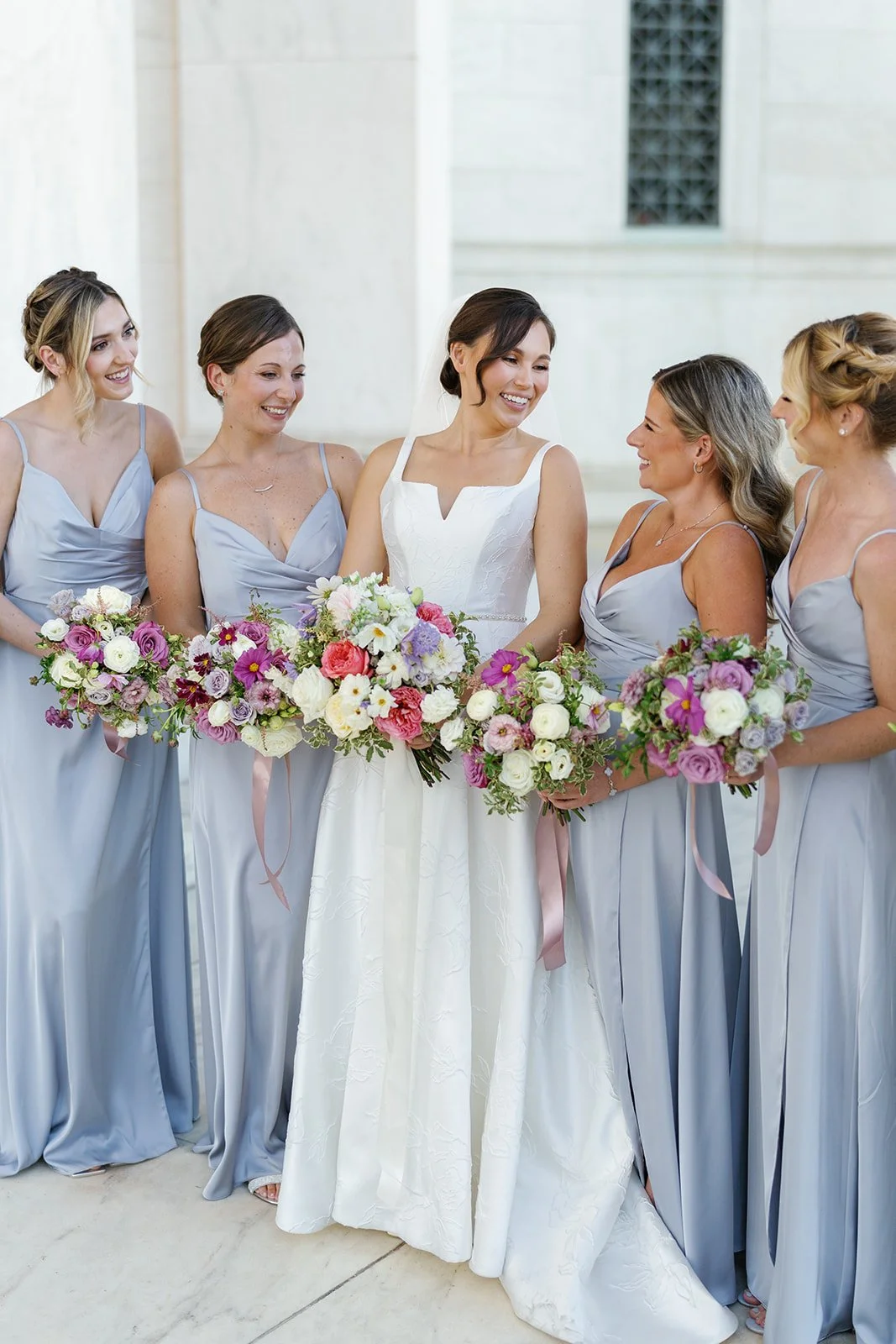 Bride surrounded by five bridesmaids holding bouquets, all wearing pastel-colored dresses, inside a light-colored venue.