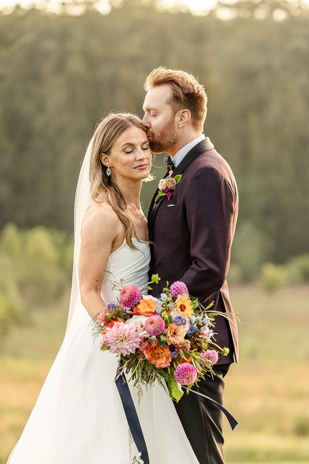 A bride and groom sharing an intimate moment outdoors, with the groom gently kissing the bride's forehead. The bride is holding a large, colorful bouquet, and both are dressed in wedding attire. The background is blurred greenery, indicating a natura