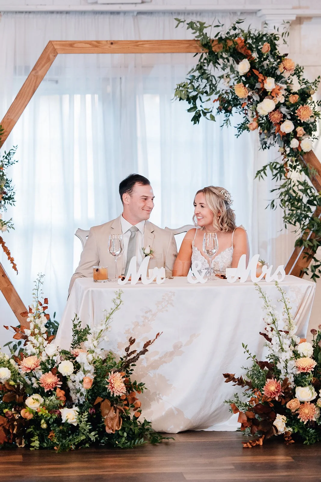 Bride and groom sit at a wedding reception table, smiling at each other, with a floral arrangement backdrop and table signs reading "Mr" and "Mrs."