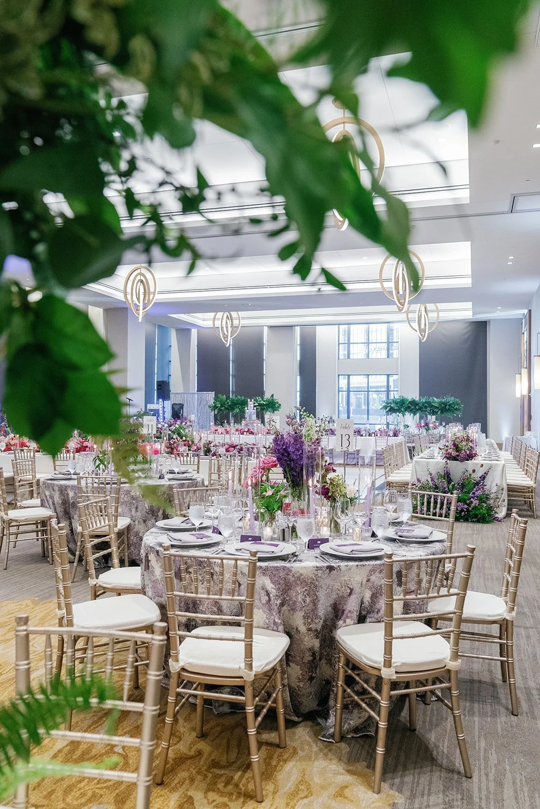 Elegant banquet hall decorated with round tables set for a formal event, featuring floral centerpieces, gold chairs, and modern lighting fixtures, with large windows allowing natural light.