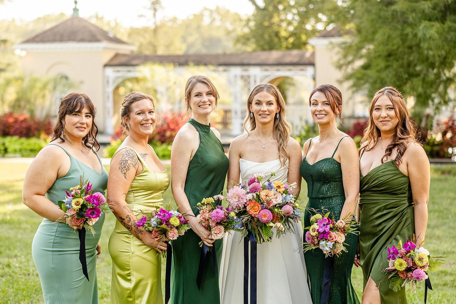 Group of seven women in colorful dresses holding bouquets of flowers, standing outdoors in a garden with a pavilion in the background.
