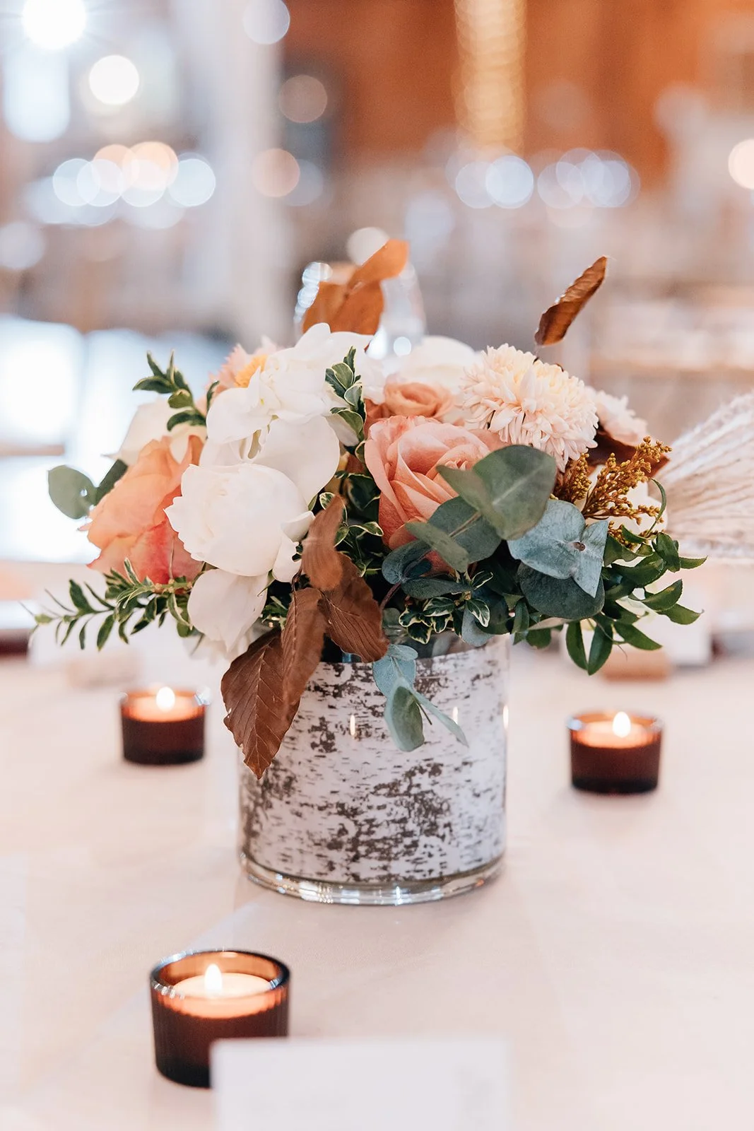 A floral centerpiece with pink and white flowers, green, brown, and gray leaves in a rustic glass vase, surrounded by small lit candles on a table.