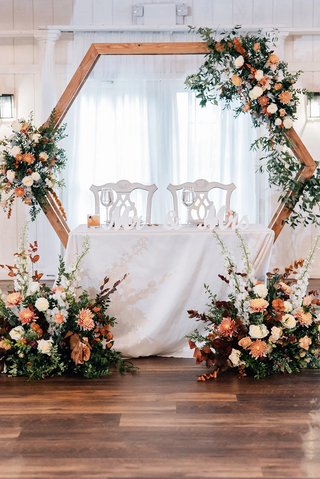 A wedding reception set-up with a wooden hexagonal arch decorated with flowers and greenery, positioned behind a white table with two ornate white chairs. The table has wine glasses and a sign that reads 'Mr. & Mrs.'. Large floral arrangements sit on