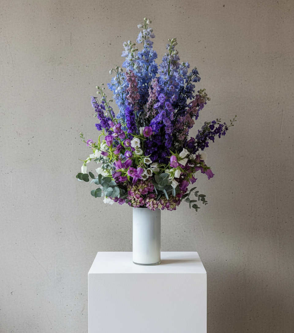 A tall flower arrangement in a white vase. It includes purple, blue, and white flowers with greenery, displayed in a room with a window.