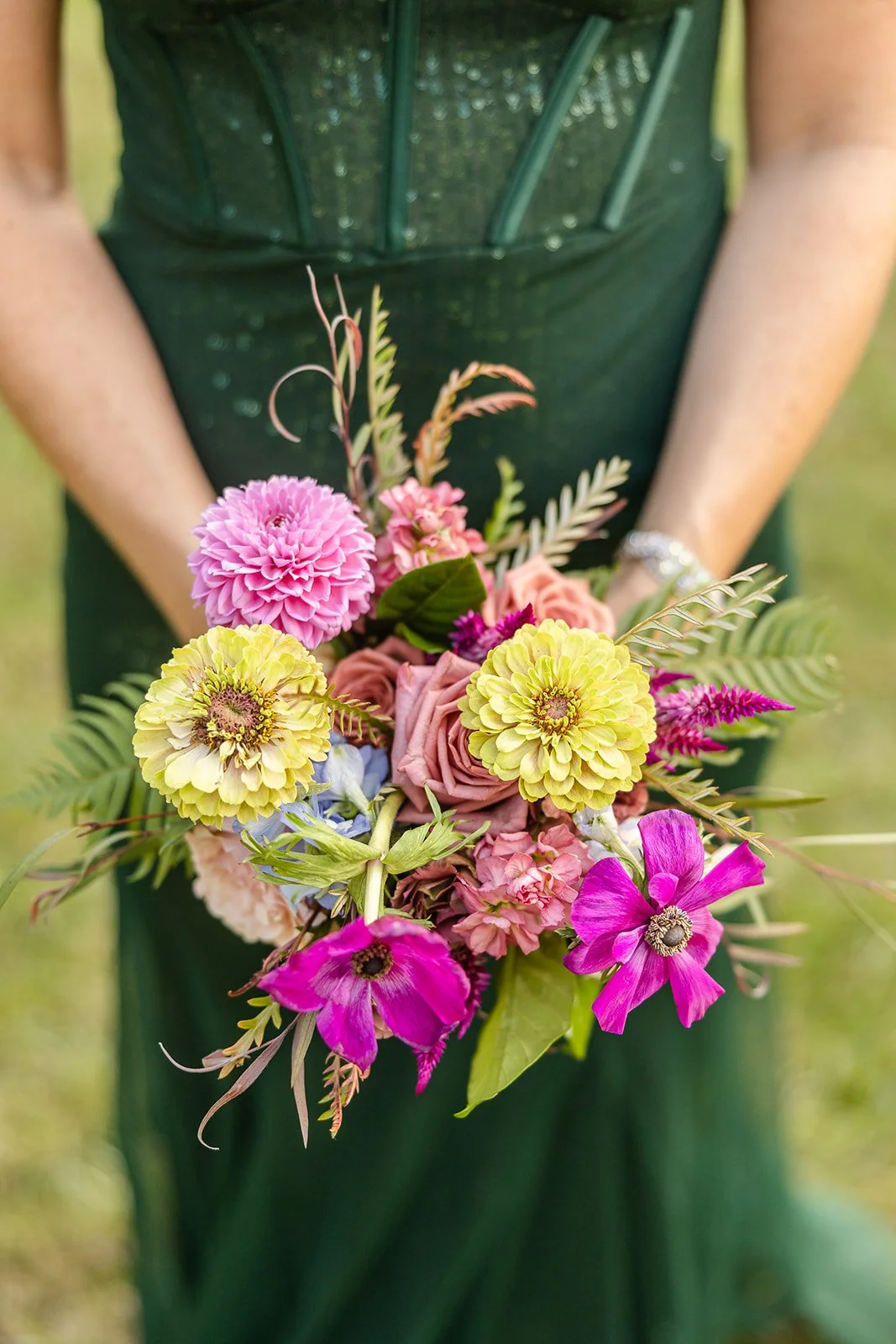 A person holding a colorful bouquet of pink, yellow, purple, and peach flowers, with green foliage, in an outdoor setting.