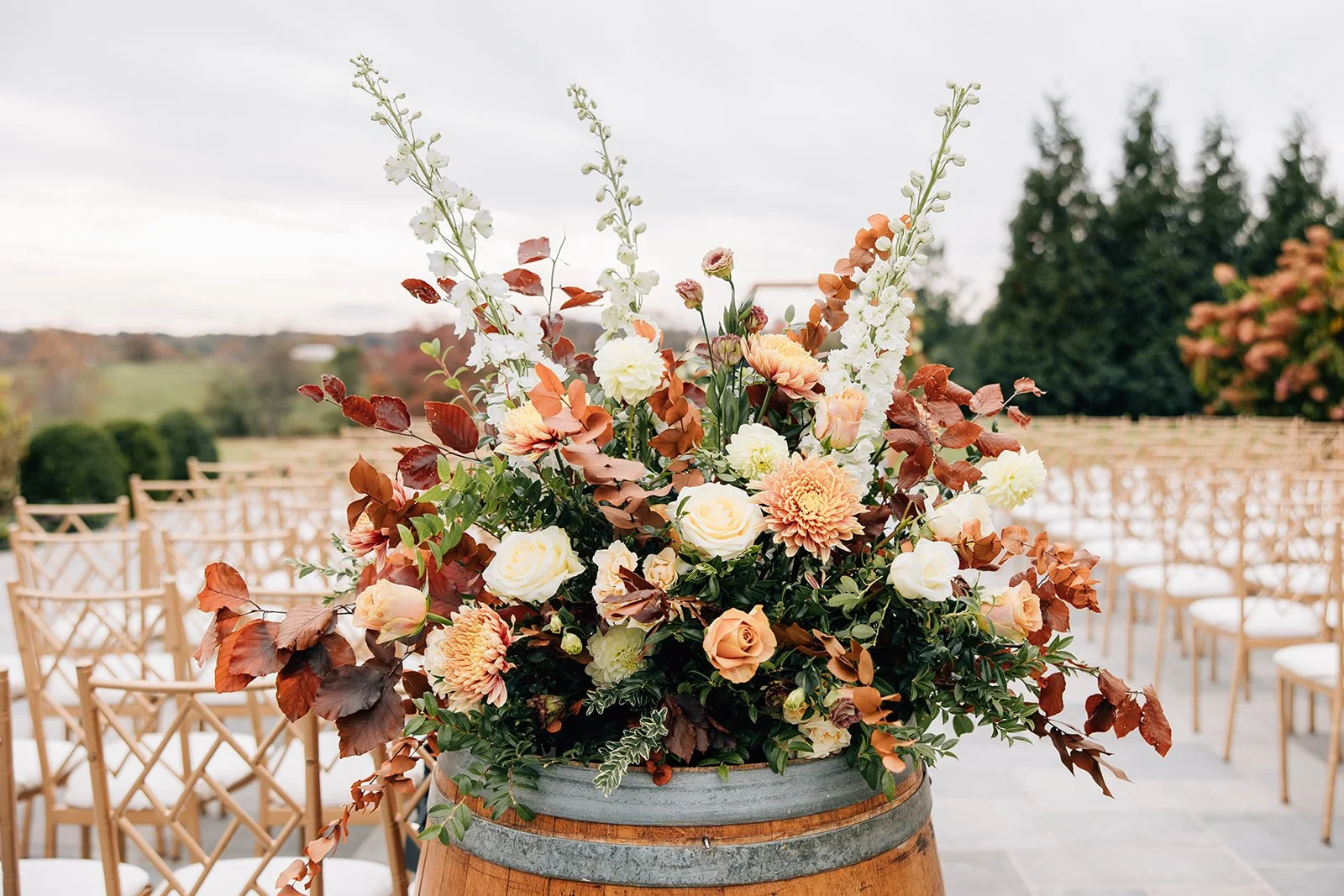Large outdoor floral arrangement with white, peach, and orange flowers, and greenery, on a barrel at a wedding ceremony site with rows of wooden chairs and a scenic landscape in the background.