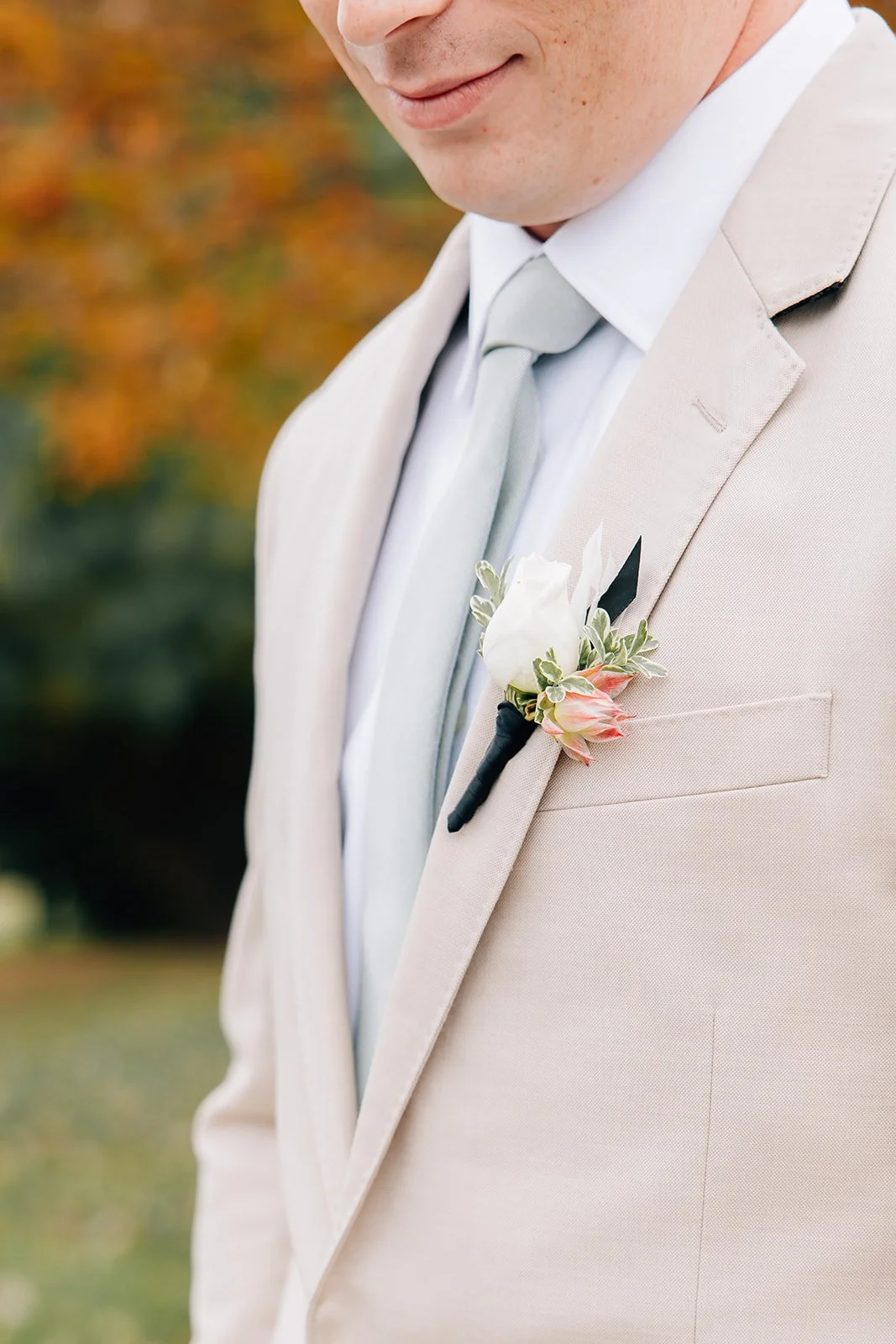 Close-up of a man in a white dress shirt, beige suit, and light gray tie, with a boutonniere of white and pink flowers on his lapel, outdoors with autumn foliage in the background.