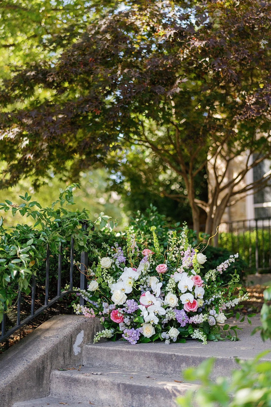A floral arrangement with white, pink, and purple flowers placed on concrete stairs at the entrance of a house, with green bushes and trees in the background.