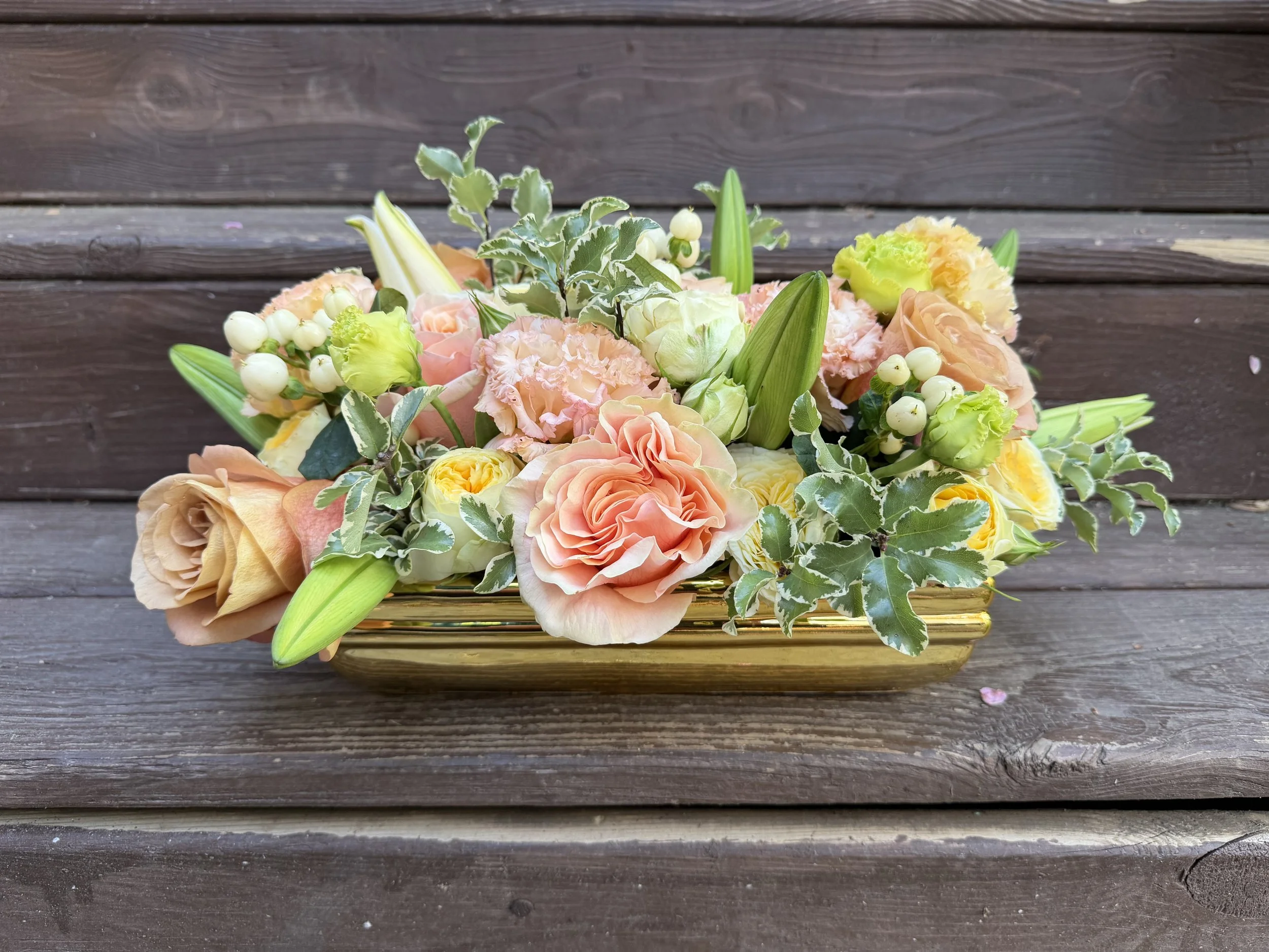 A bouquet of peach, cream, and white flowers with green leaves in a gold striped vase, placed on a wooden surface.