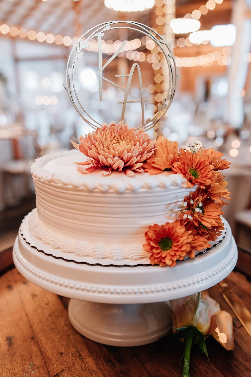 A white wedding cake decorated with orange flowers and topped with a personalized cake topper that reads 'K + A' with a circular border. The cake is on a white stand with a wood background and warm string lights.