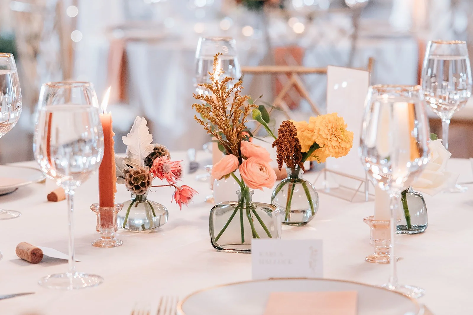 Elegant table centerpiece with small glass vases holding pink and yellow flowers, a lit peach-colored candle, and wrapped corks in a formal dining setting.