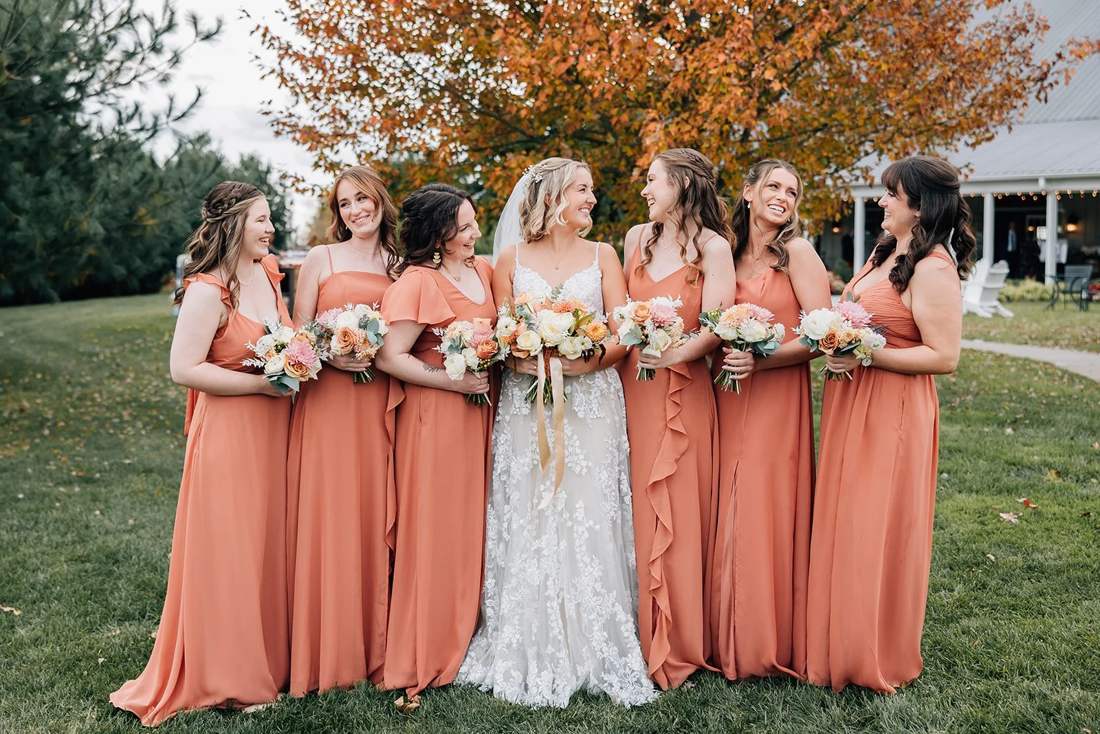 A bride and her six bridesmaids standing on a grassy lawn outdoors, dressed in matching burnt orange bridesmaid dresses, holding bouquets of flowers, with autumn trees and a building with string lights in the background.