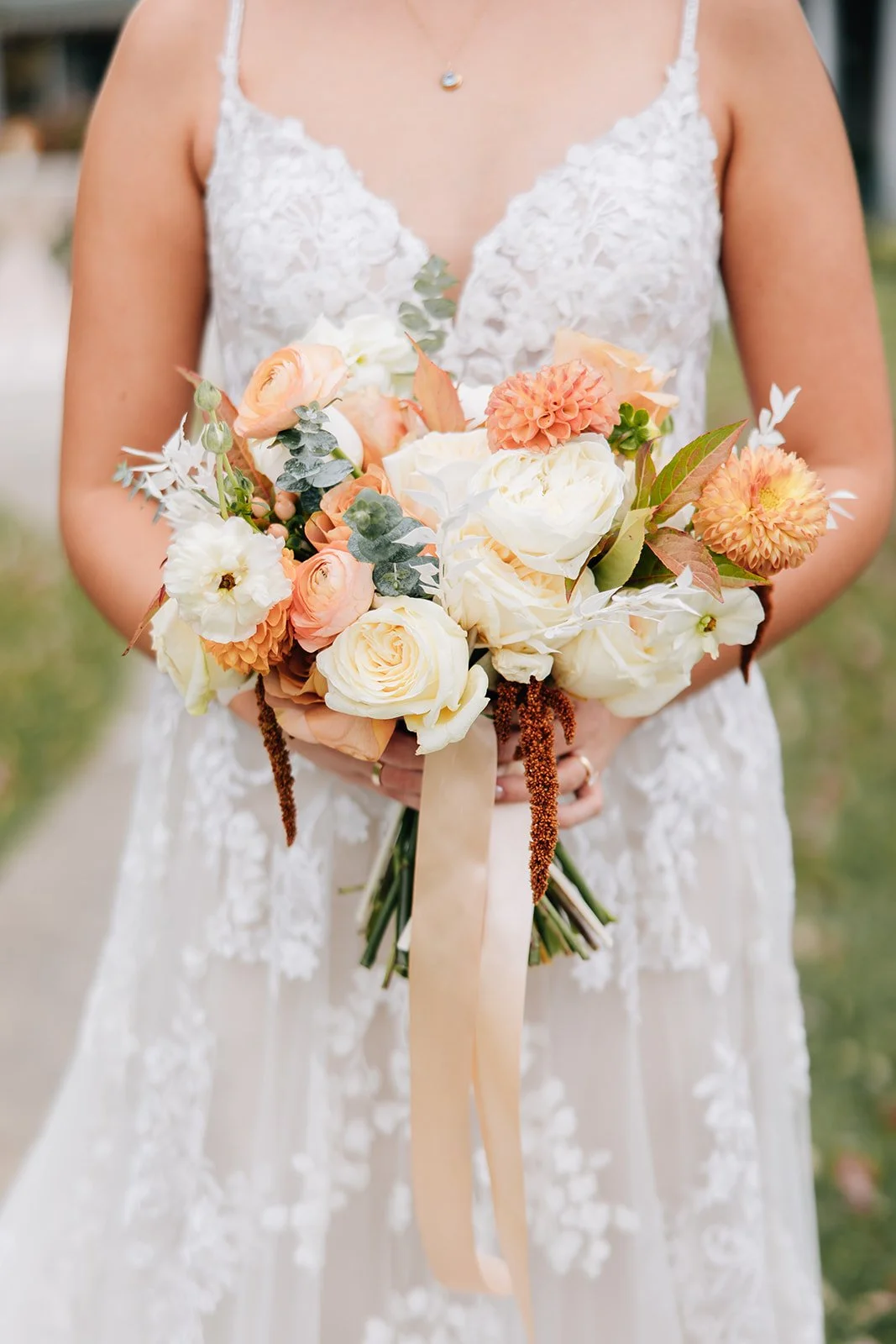 A bride in a lace wedding dress holding a bouquet of cream, peach, and orange flowers, including roses and dahlias, with greenery and a beige ribbon.