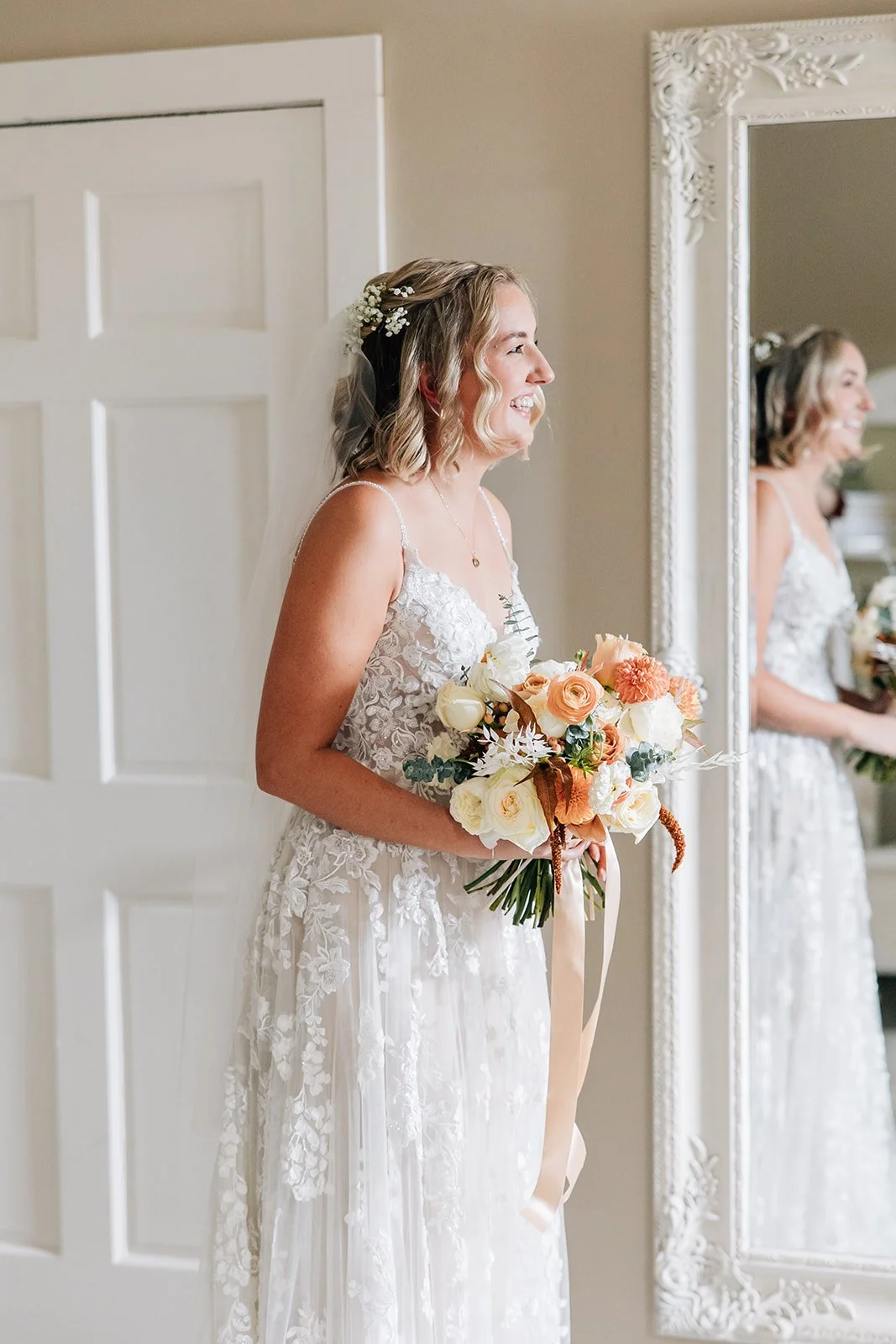 A bride with wavy blonde hair, holding a bouquet of white and peach flowers, standing next to a mirror and smiling.