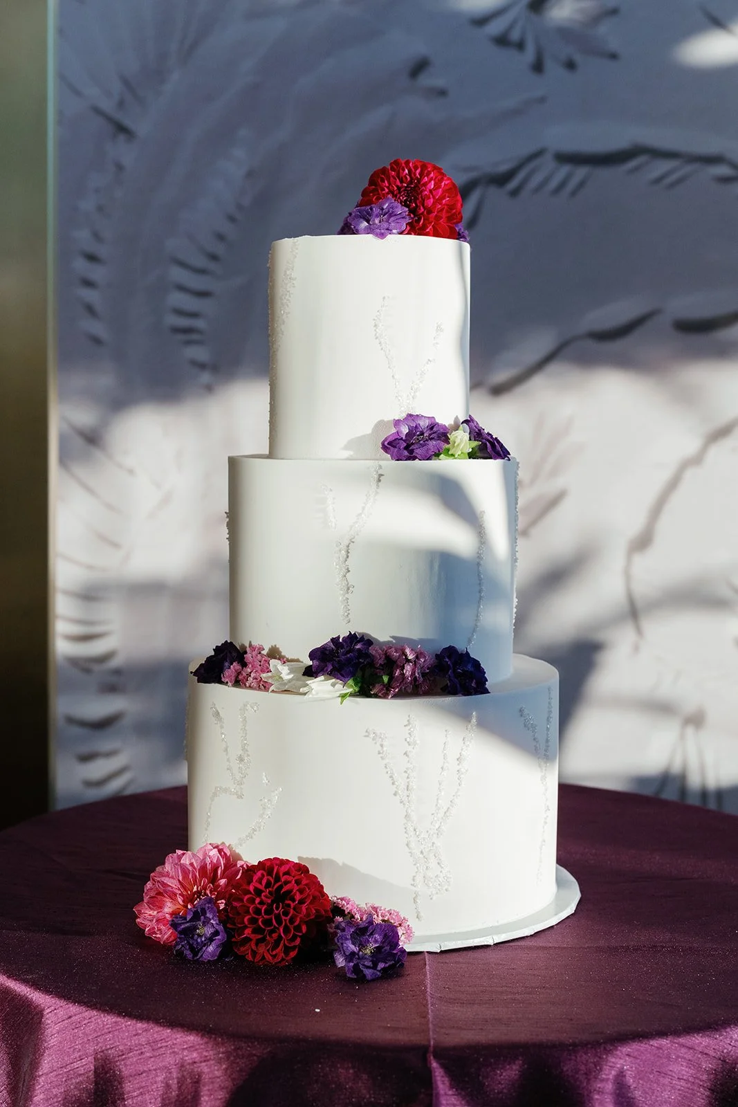 Multi-tiered white wedding cake decorated with purple and red flowers, placed on a purple tablecloth.