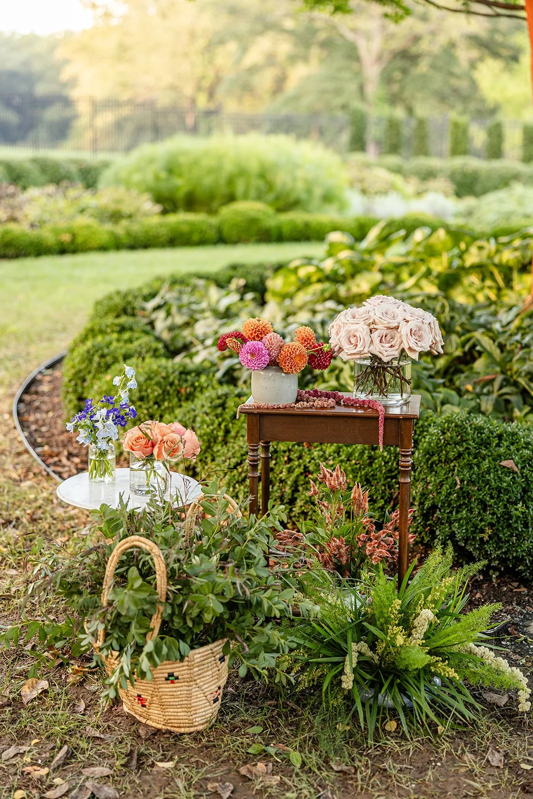 Outdoor garden scene with various flower arrangements on small tables and baskets, featuring roses, dahlias, and irises, surrounded by greenery and lawn.