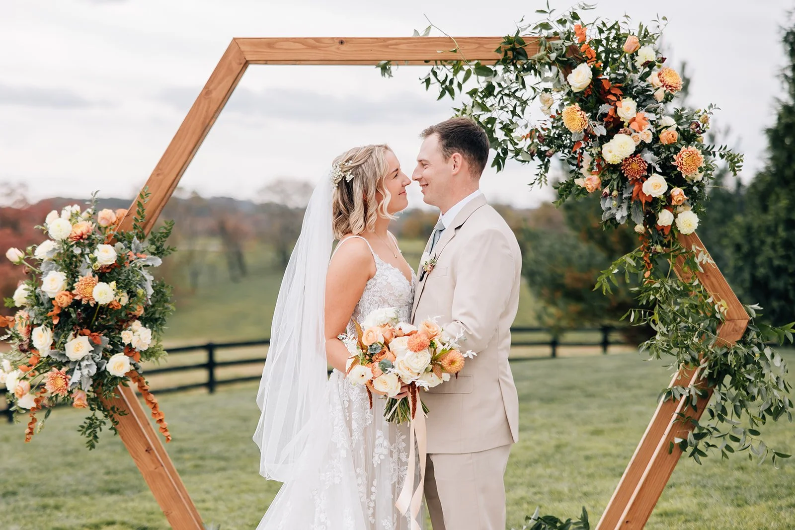 A bride and groom standing close, facing each other, smiling, in front of a wooden wedding arch decorated with flowers, outdoors on a cloudy day.