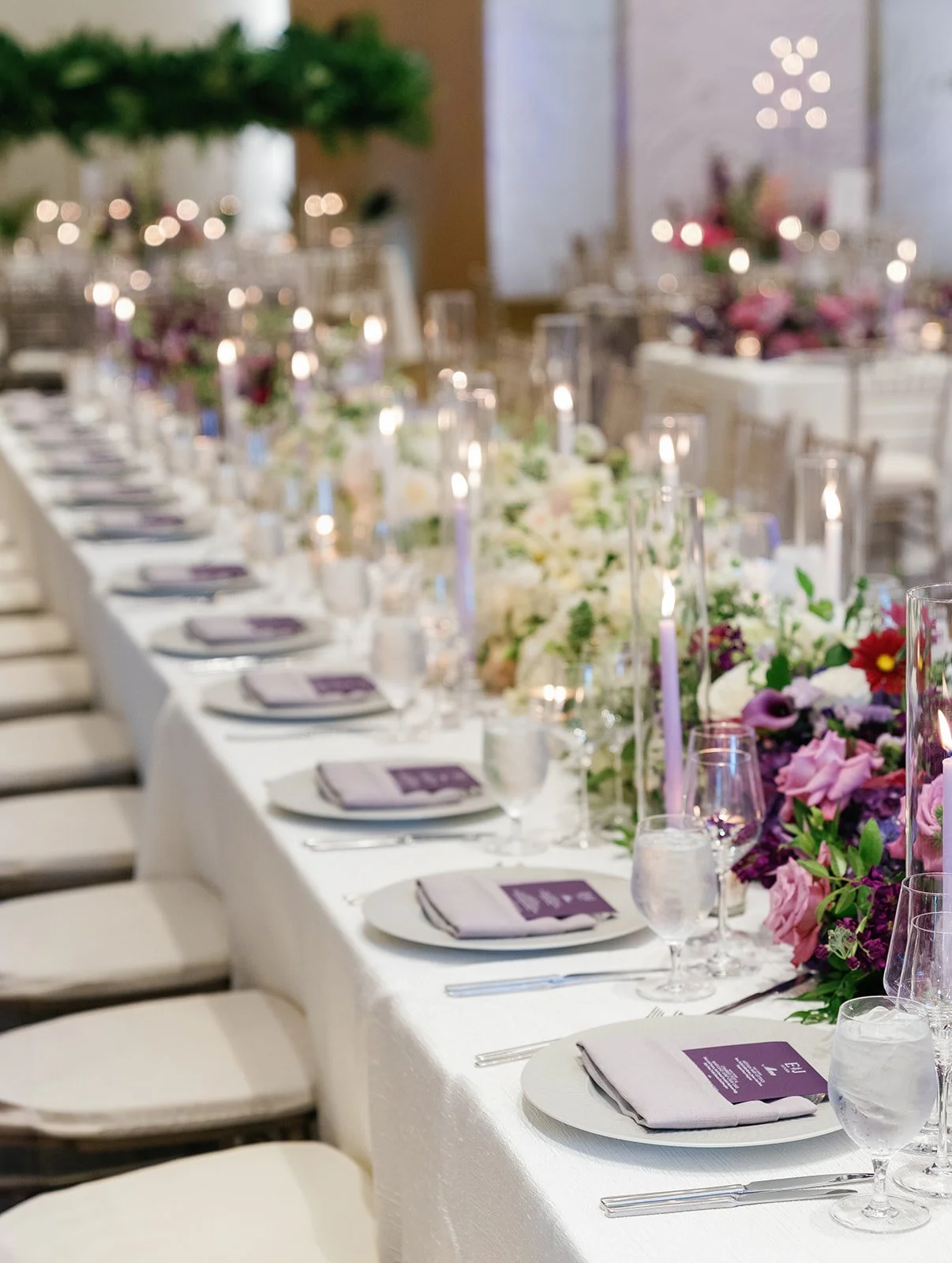Elegant banquet table set with white tablecloth, purple napkins, clear glassware, silverware, and a floral centerpiece with pink, white, and purple flowers, in a decorated event hall.