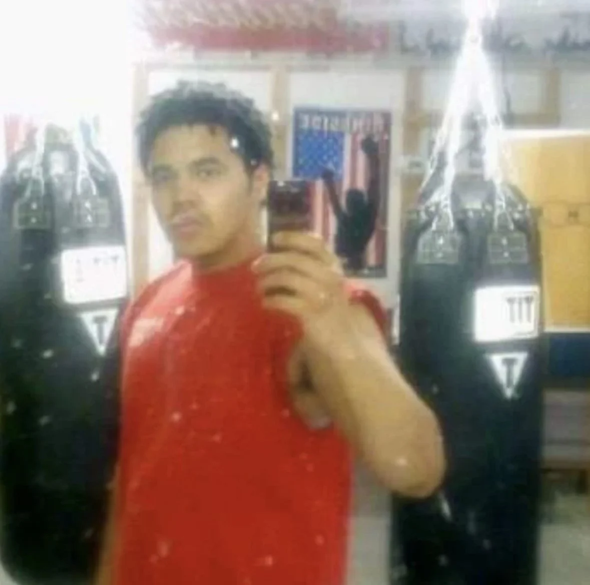 A young man taking a mirror selfie in an indoor space, with two large bottled drinks and a person hanging clothes in the background.