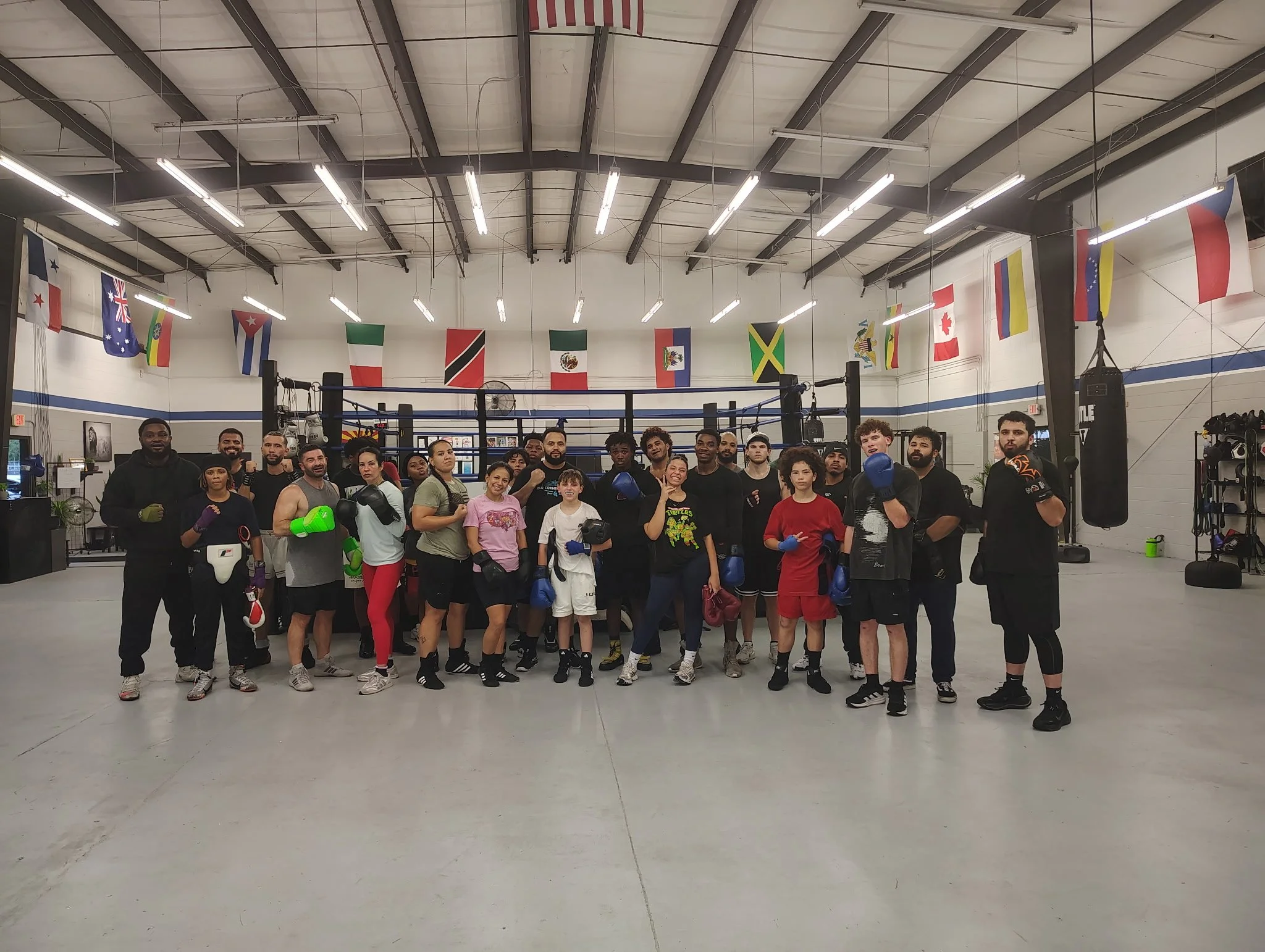 Group of diverse people posing in a boxing gym with boxing equipment and flags hanging from the ceiling.
