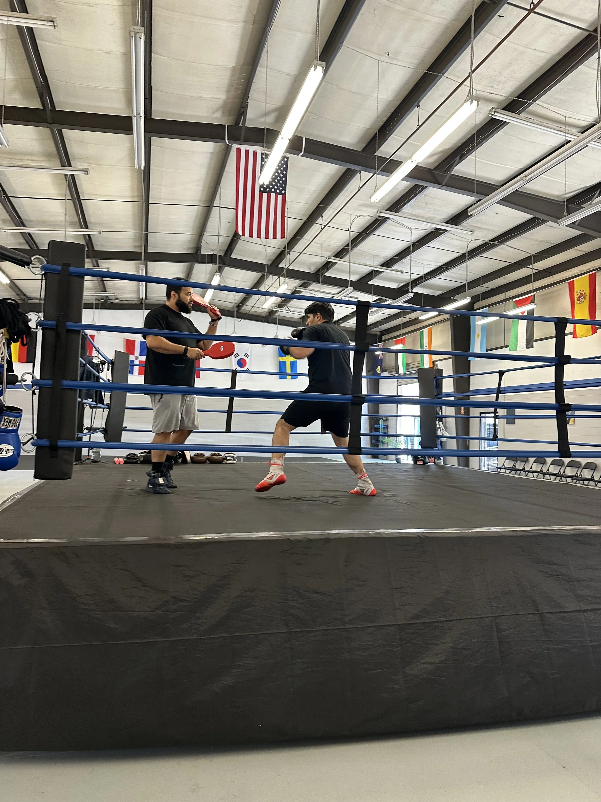 Two men are boxing inside a boxing ring in a gym with flags of different countries hanging from the ceiling.