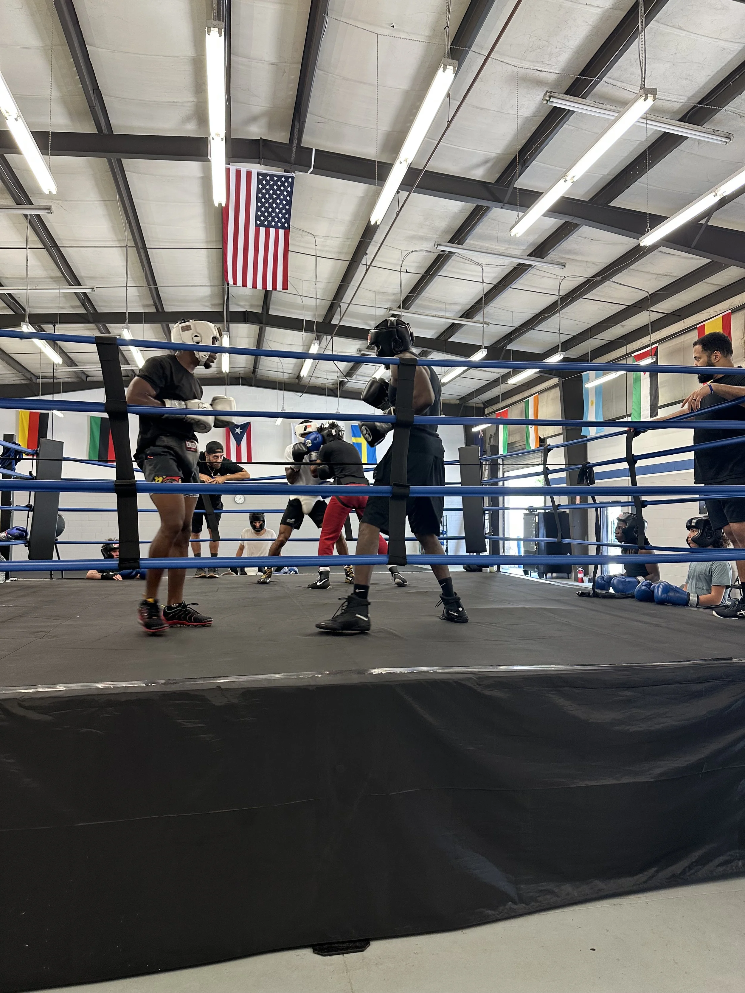 Two boxers in black shorts and shirts sparring in a boxing ring inside a gym. One boxer is wearing a white headgear, and the other is wearing black headgear. Several people are watching, some seated and some standing, and various flags hang from the ceiling.