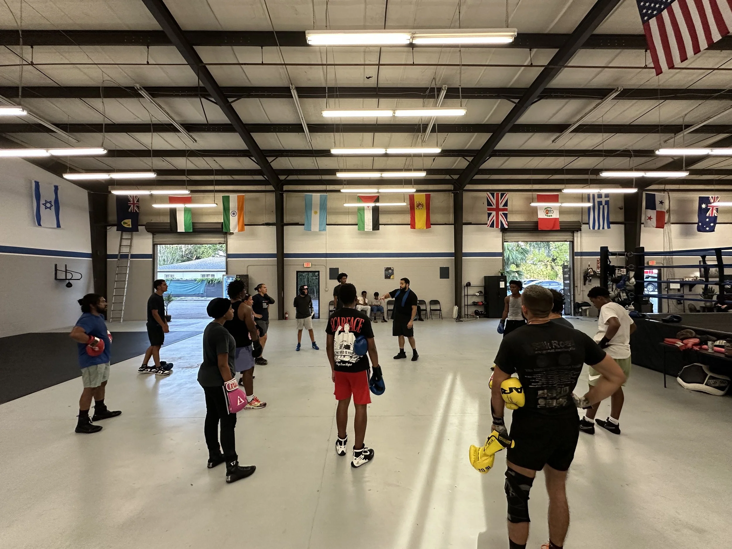 A group of people gathered inside a boxing gym, holding boxing gloves, standing in a circle, appearing to listen to a trainer or instructor. The gym has flags from various countries hanging from the ceiling and some workout equipment on the sides.