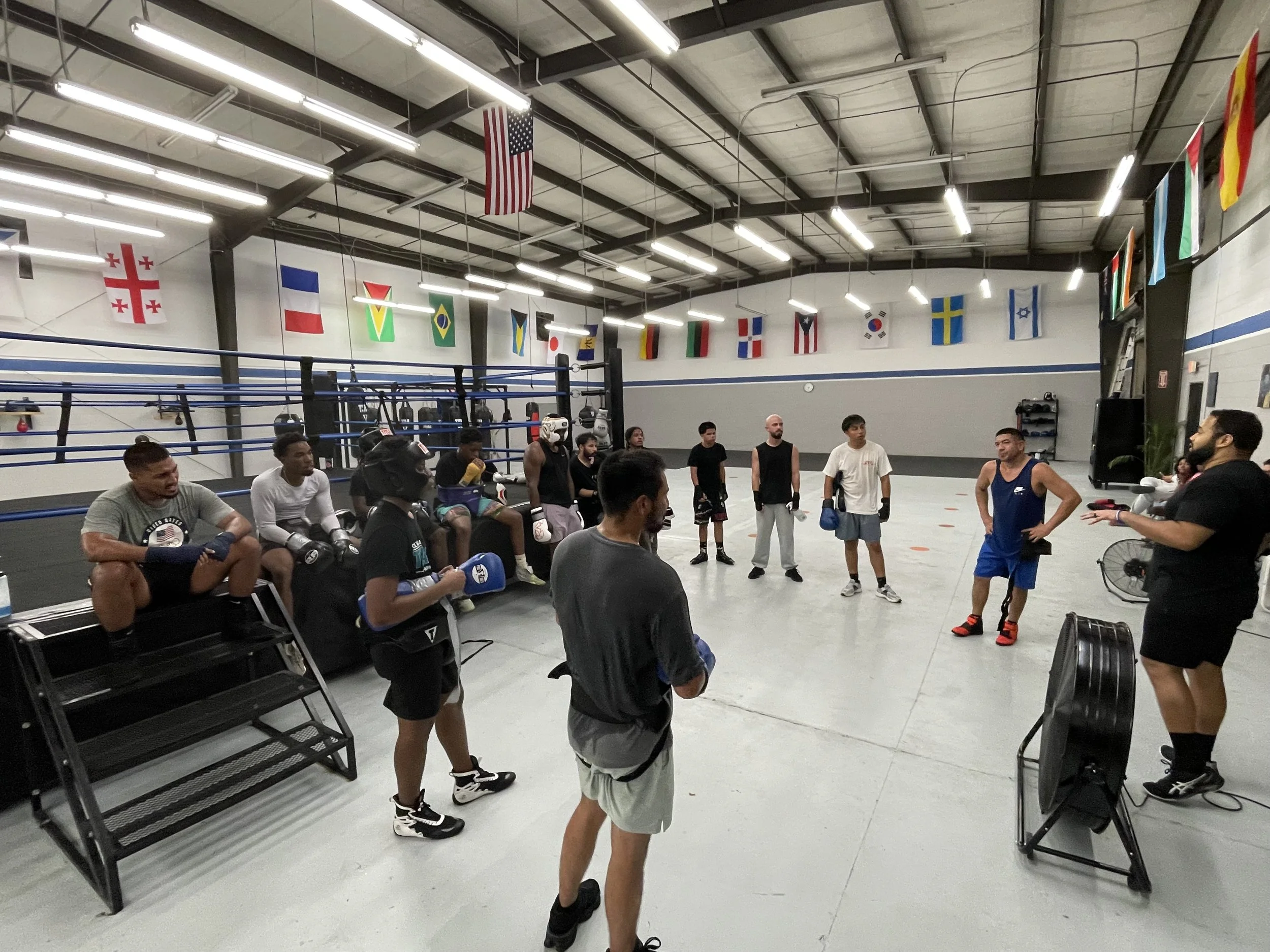 Boxers and trainers gather in a gym with international flags hanging from the ceiling, some seated and others standing while discussing or preparing for a training session.