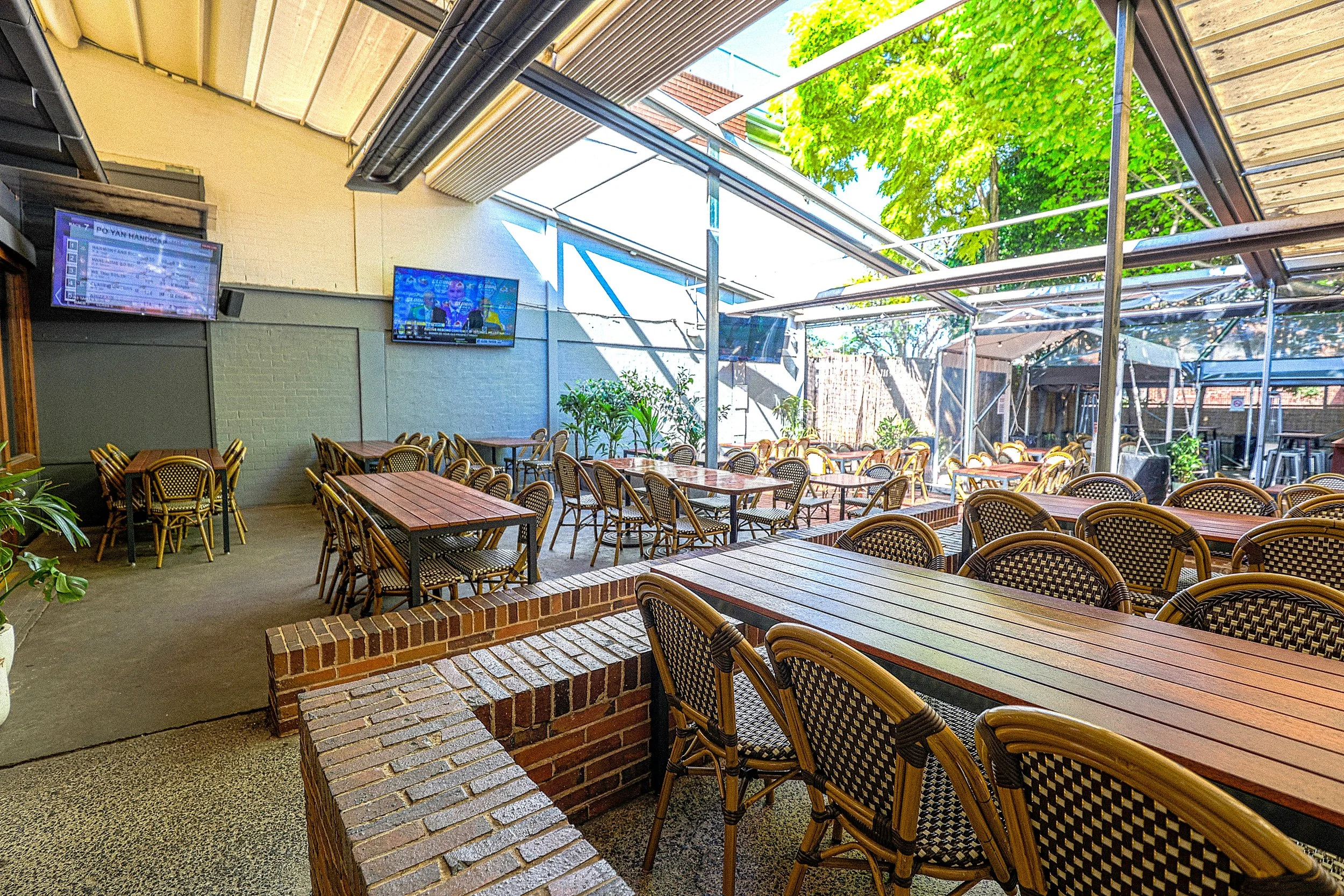 Empty outdoor restaurant patio with wooden tables and woven chairs, partially covered with a roof, surrounded by green trees and potted plants, with TV screens mounted on the wall.