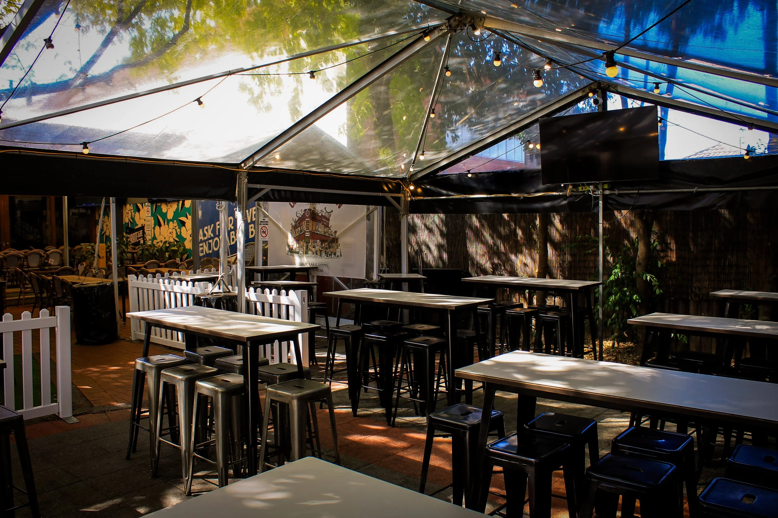 Empty outdoor patio with high tables and bar stools under a blue canopy and string lights, surrounded by greenery and a fence.