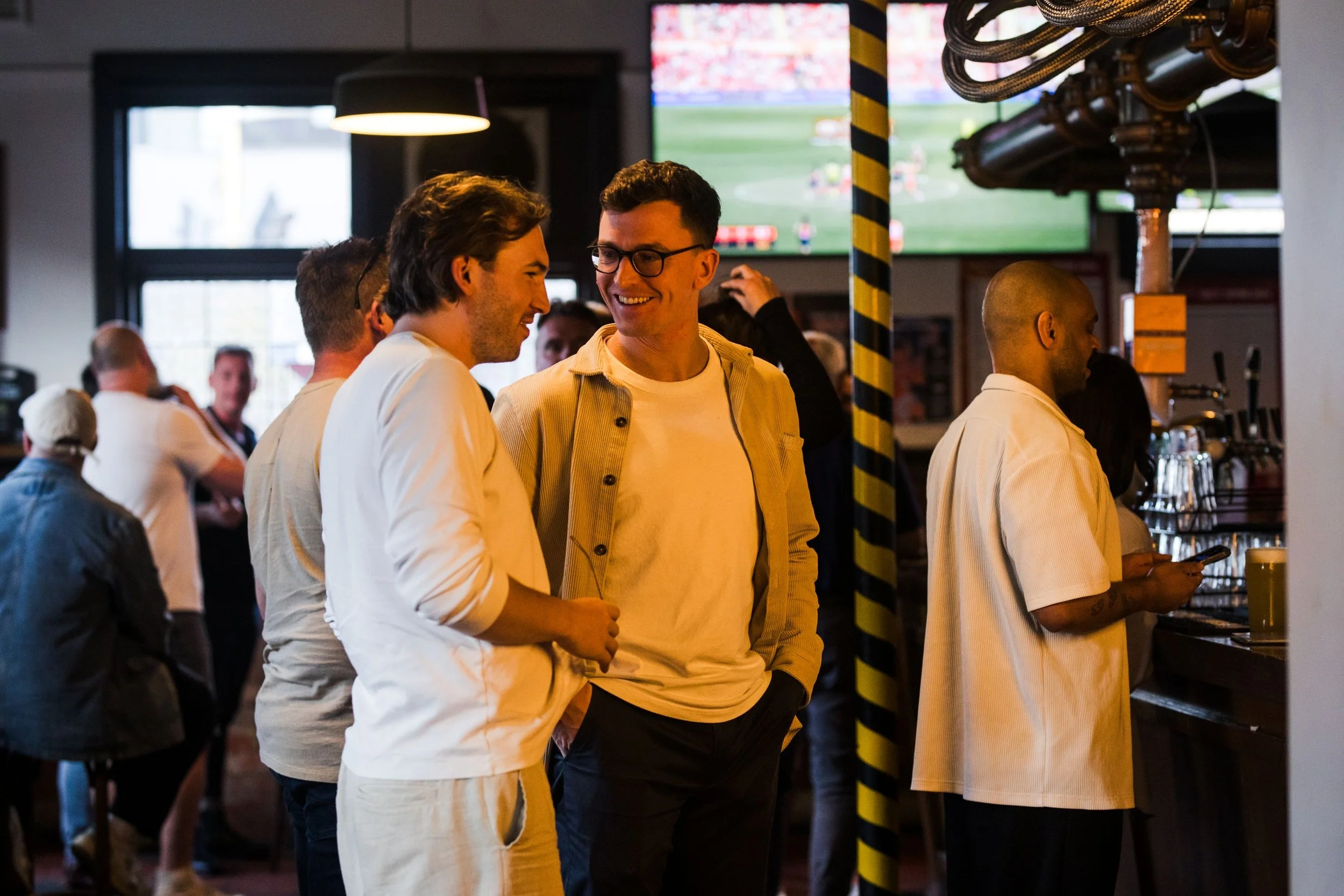People socializing at a bar or pub, including two men smiling and talking near the bar counter, with others in the background and a television screen showing a sports game.