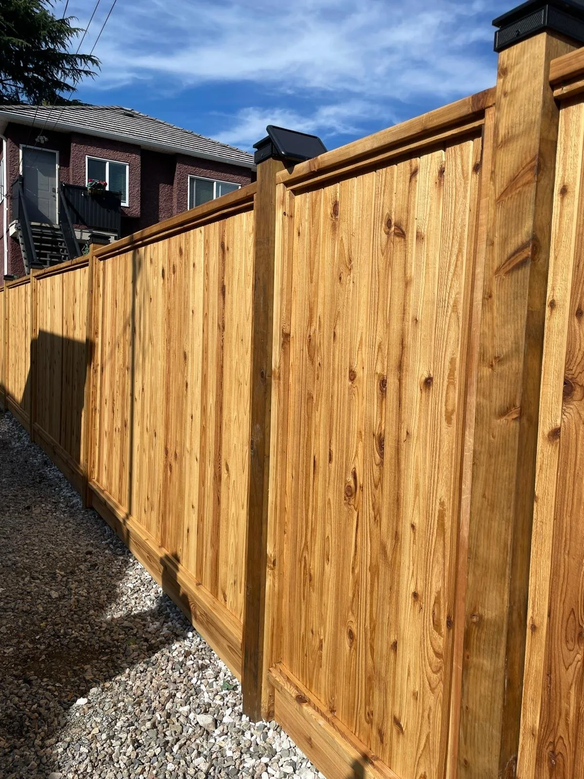 Newly installed wooden privacy fence with vertical planks and supporting posts, under a partly cloudy sky, with a residential building in the background.