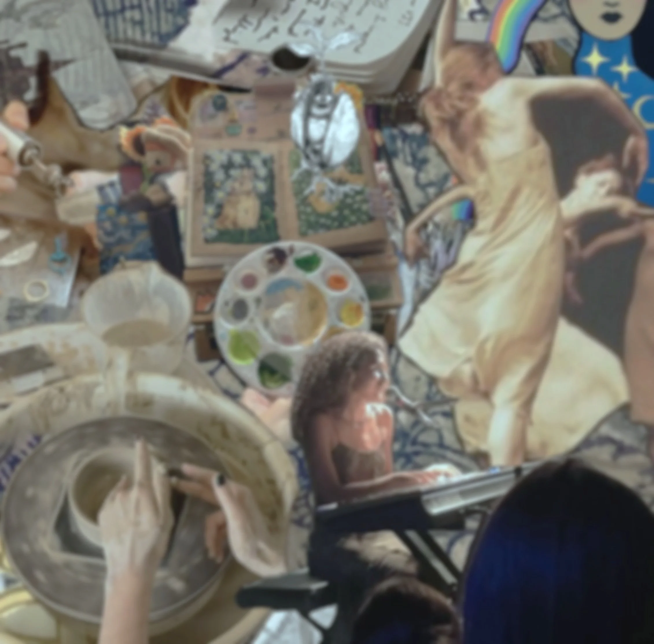 A messy table with art supplies, an open sketchbook, and a variety of painted and unpainted figurines, including a dog figure and a person figure, alongside a painter's palette with colorful paints and a hand mixing paint in a bowl.