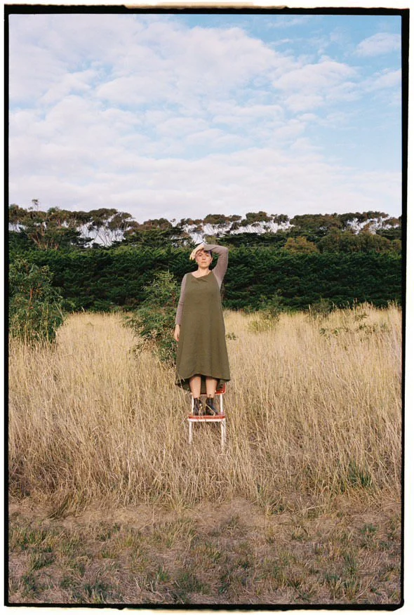 A woman standing on a chair in a field with tall yellow grass, holding her hand to her forehead, looking into the distance. Behind her are dense green trees and a partly cloudy sky.
